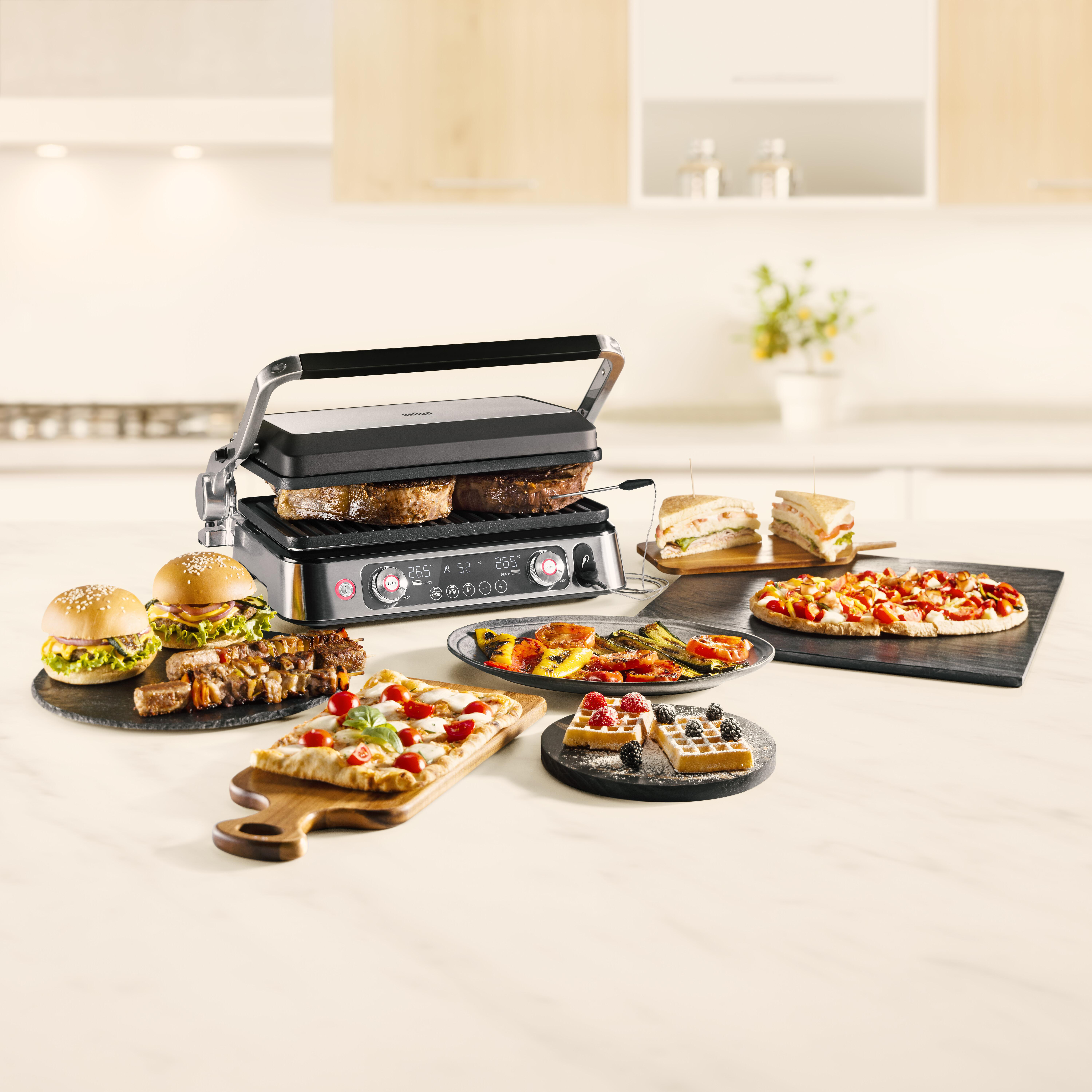 Elektrischer Grill mit gegrillten Speisen: Burger, Pizza, Waffeln und Gemüse, auf einer Küchenarbeitsplatte.