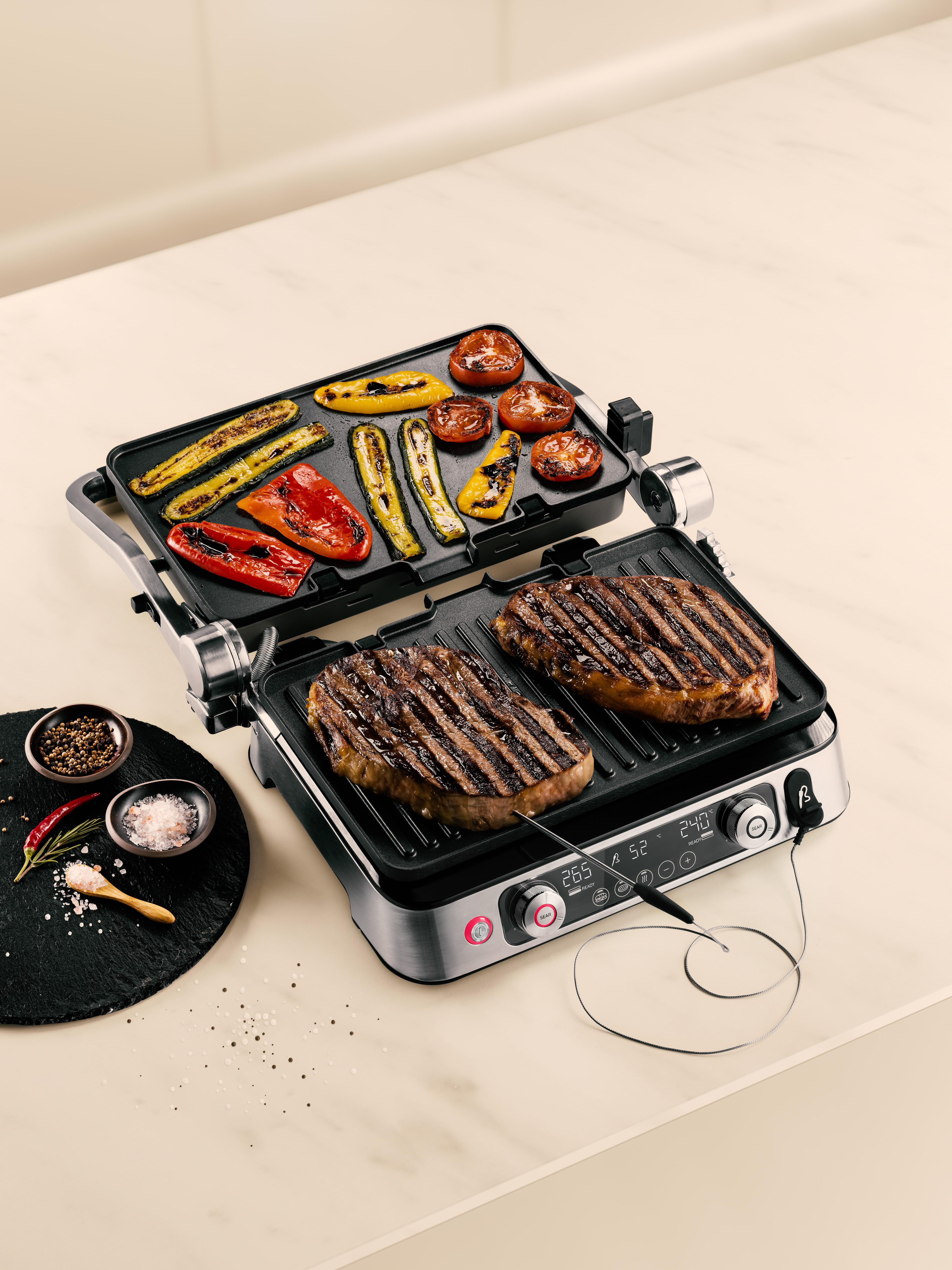 Ein Grill mit gegrilltem Gemüse und Steak auf einer weißen Oberfläche, mit Salz, Pfeffer und Chilischoten.