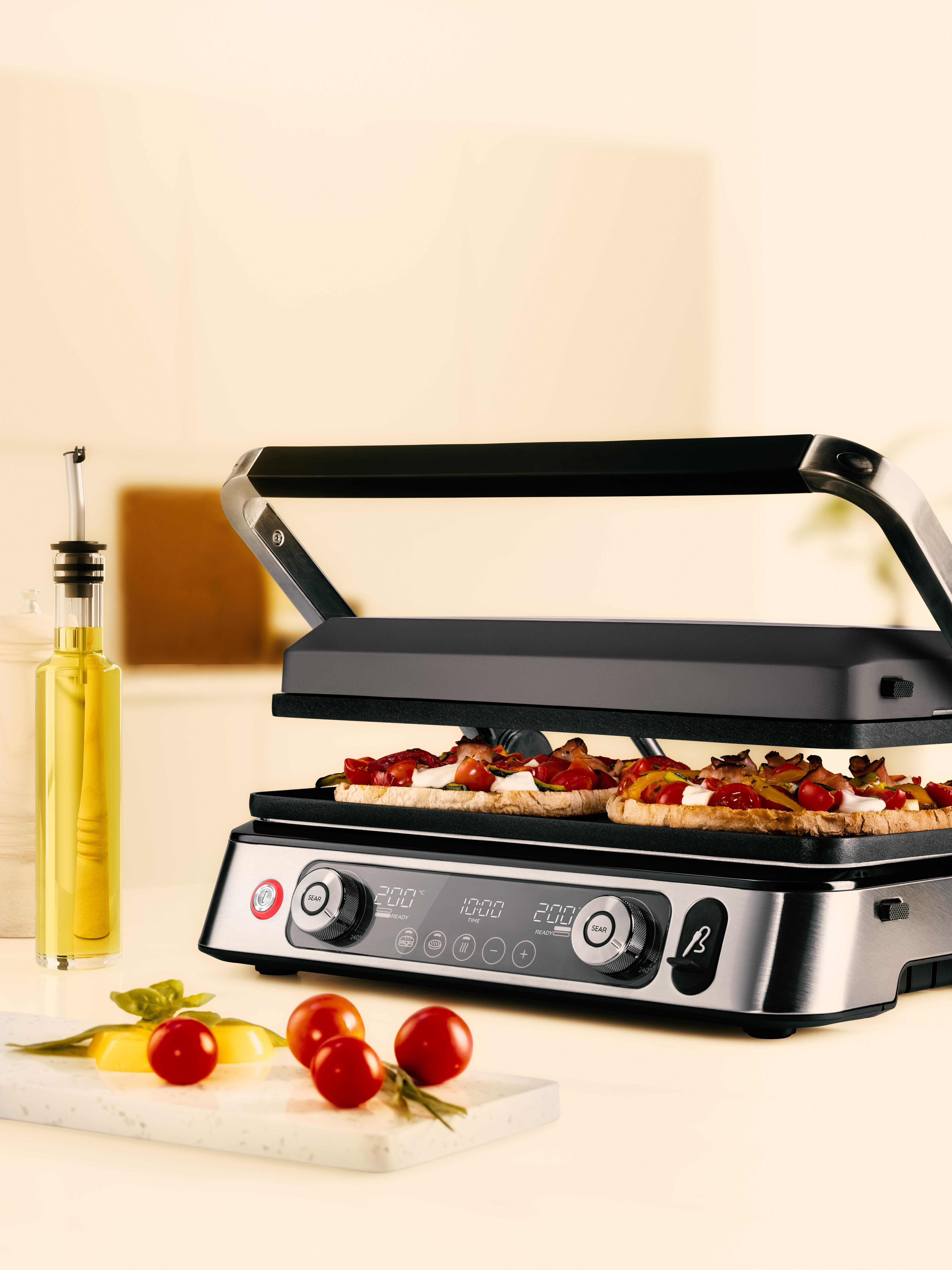 Indoor-Grill mit Essen darauf. Tomaten und Ölflasche links. Weißer Hintergrund.