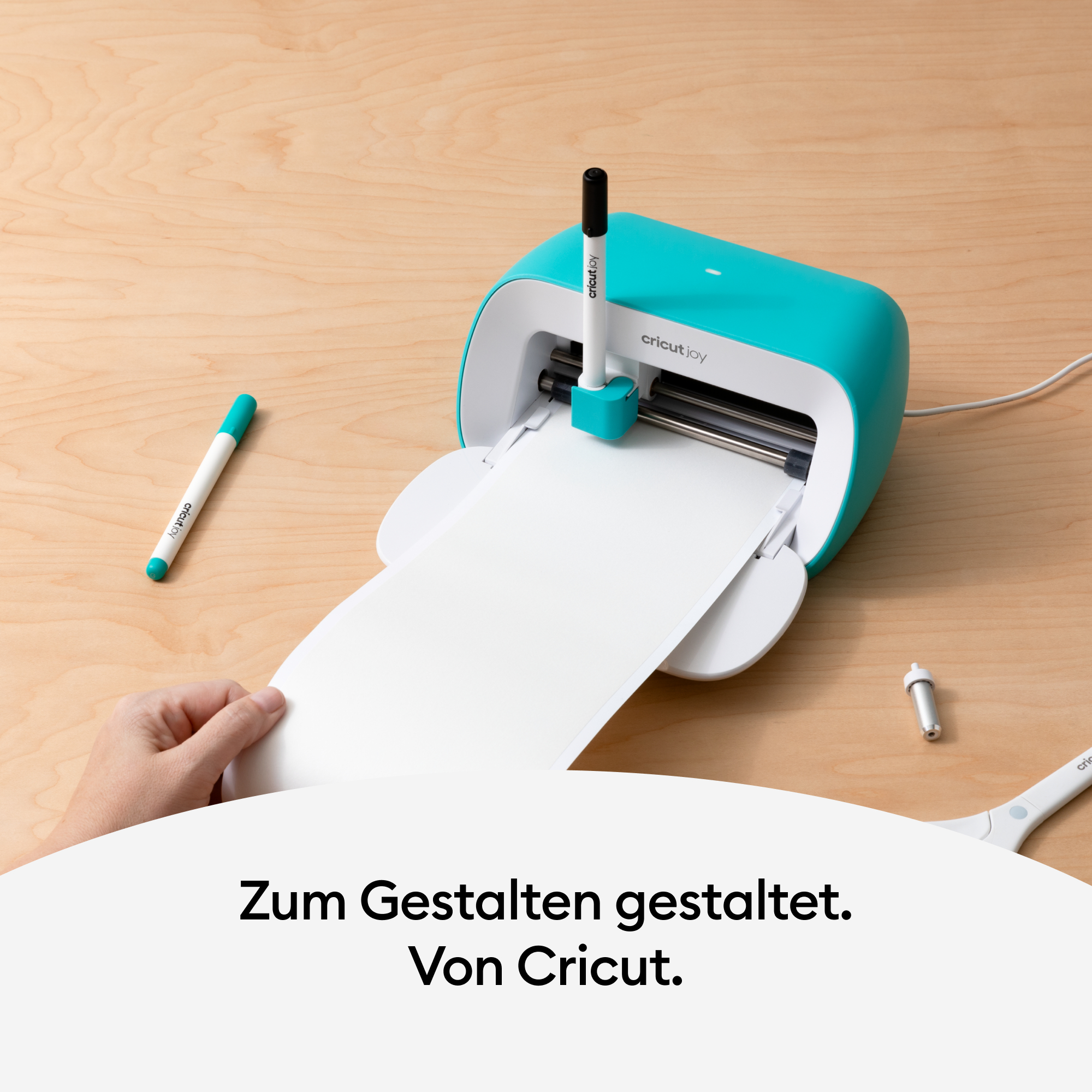 Eine türkisfarbene Cricut Joy-Maschine mit Stift und Papier. Eine Hand links.