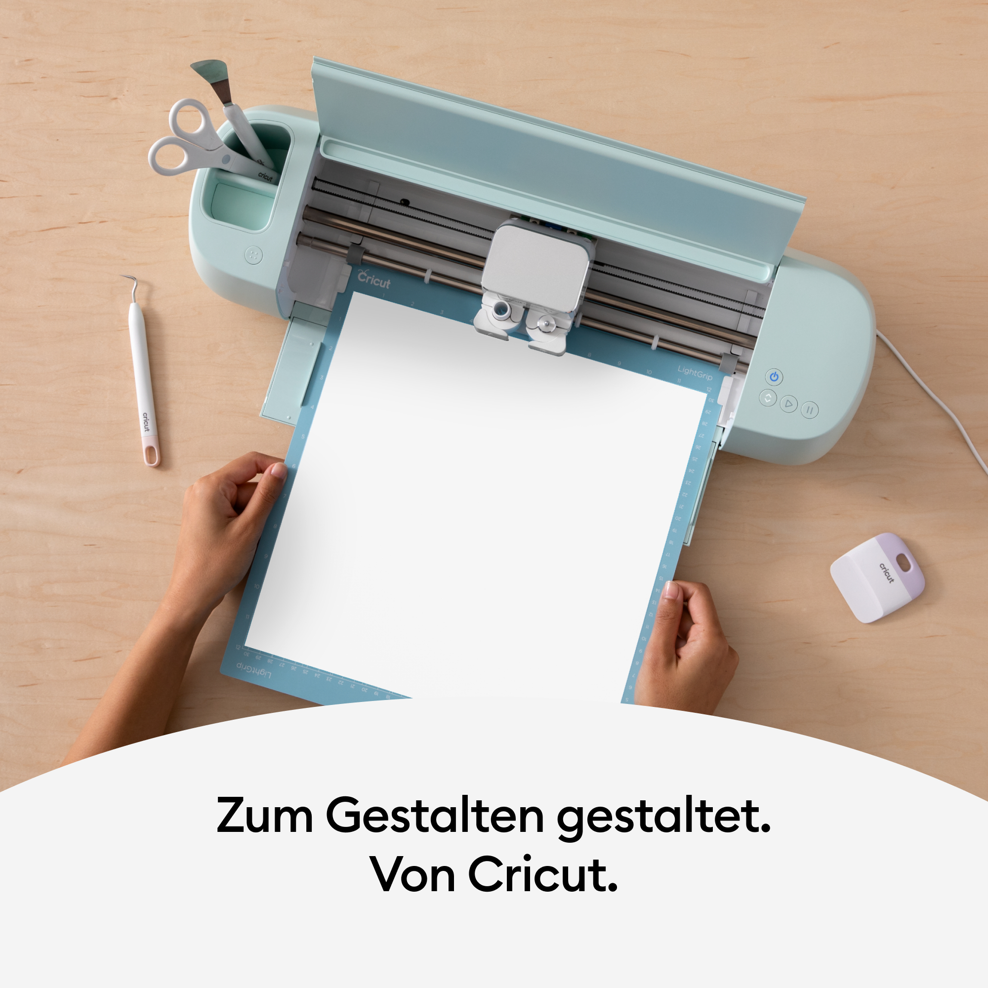 Hände halten eine blaue Schneidematte mit weißem Papier und eine Cricut-Maschine im Hintergrund.