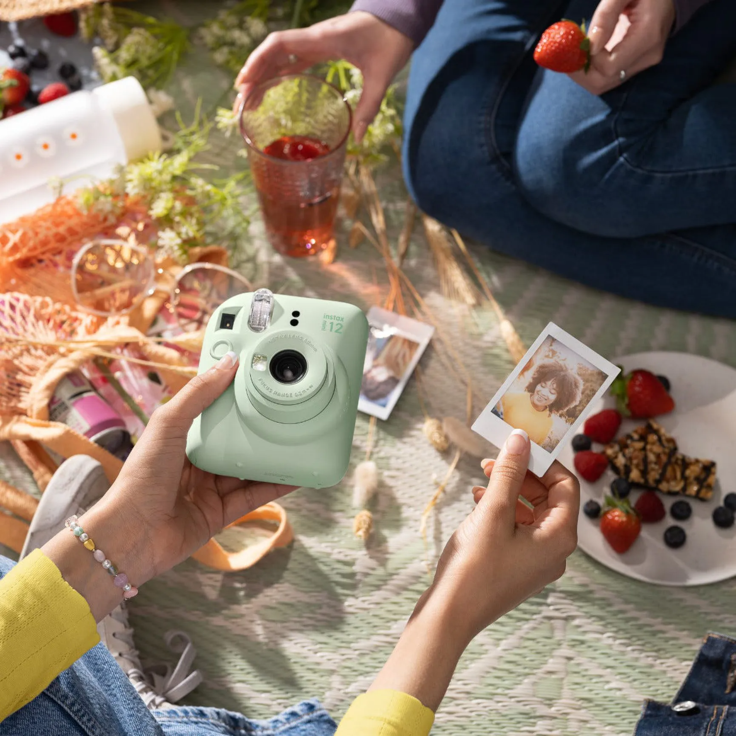 Hands holding a mint green camera, a photo, and a glass of red drink, picnic setting.