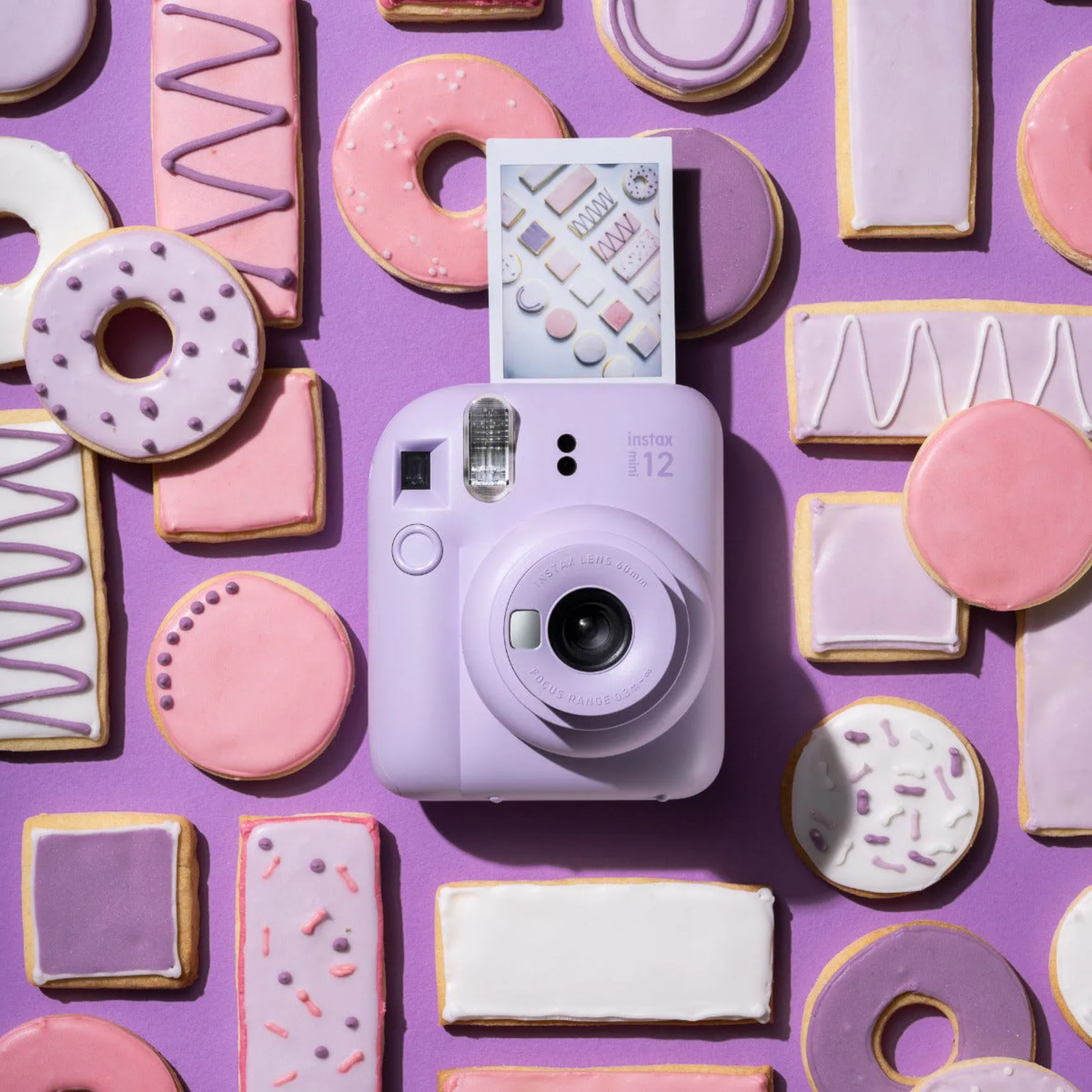 Un appareil photo Instax violet est posé parmi des biscuits et des beignets sur un fond violet. La photo montre des formes de biscuits.