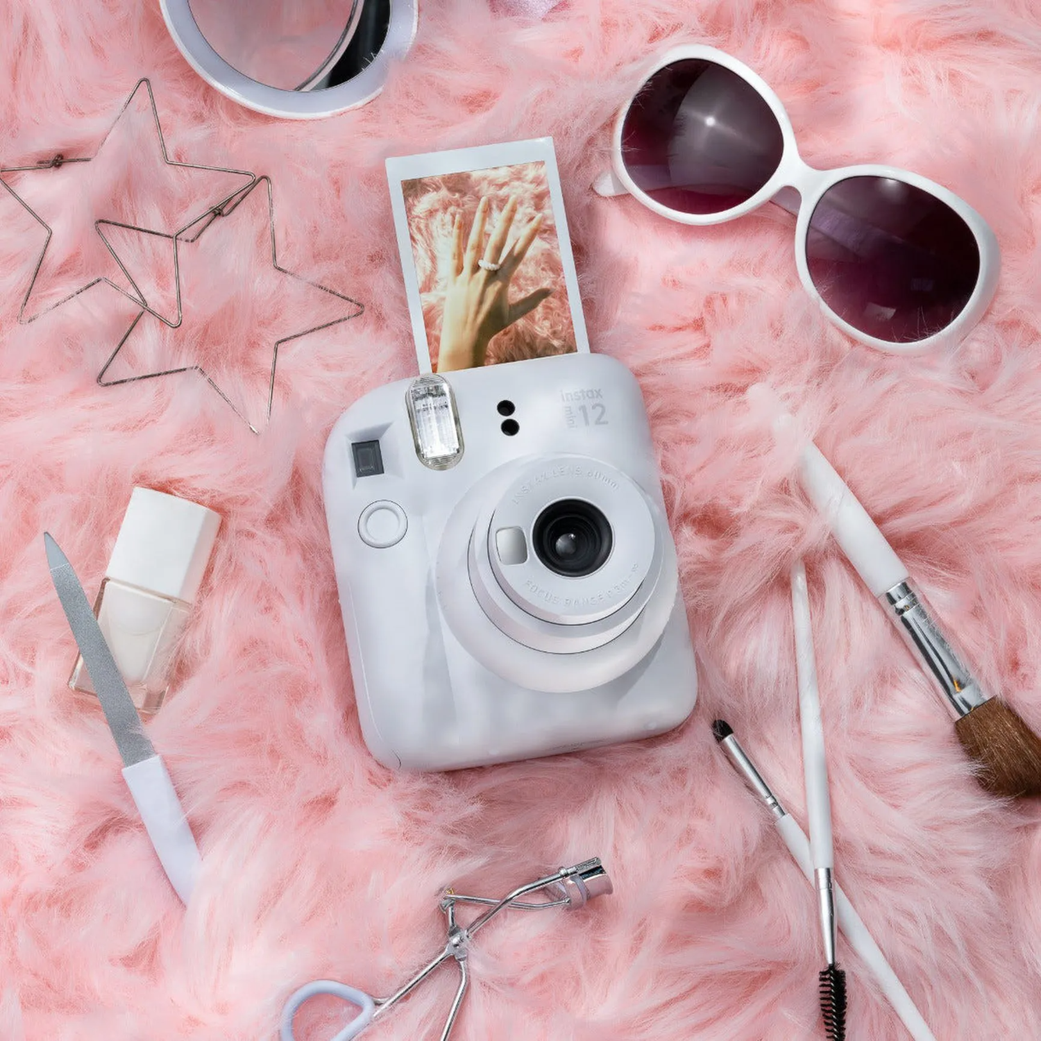 A white Instax camera surrounded by beauty products on a pink, fluffy surface. A photo is in the camera.