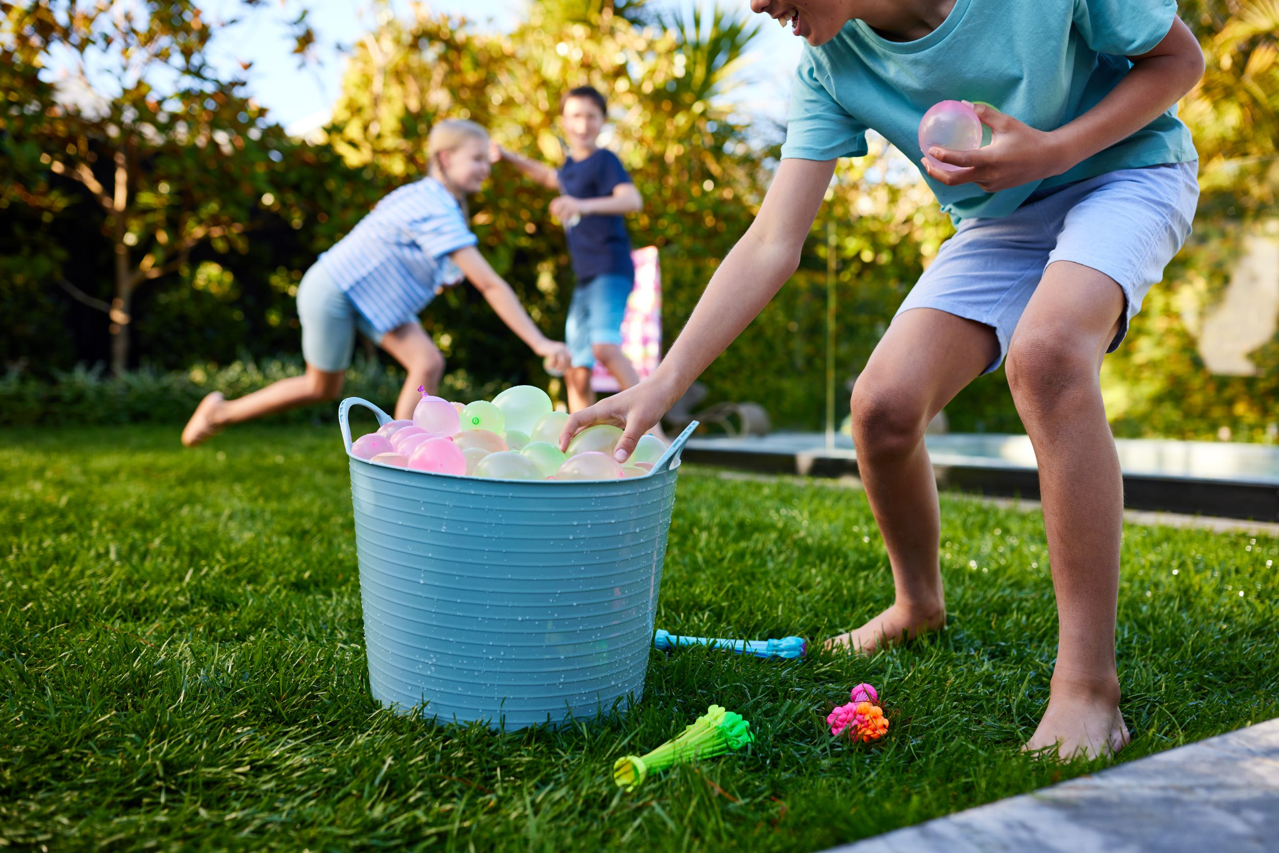 Kinder spielen Wasserspiel. Grünes Gras, blauer Eimer mit Wasserballons und ein sonniger Tag.