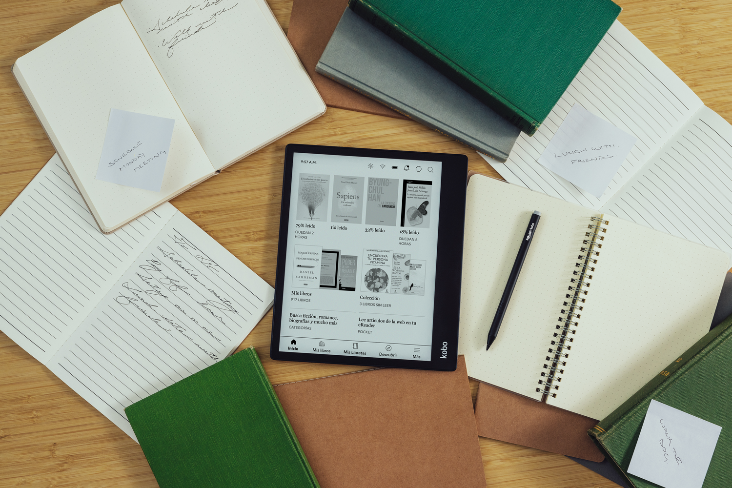 Un libro abierto, una tableta y cuadernos están sobre una mesa de madera. Hay notas adhesivas.