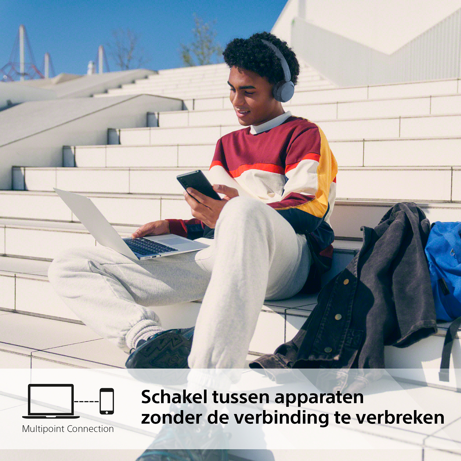 Een jonge man zit op een trap, gebruikt een laptop en houdt een telefoon vast, met een koptelefoon.