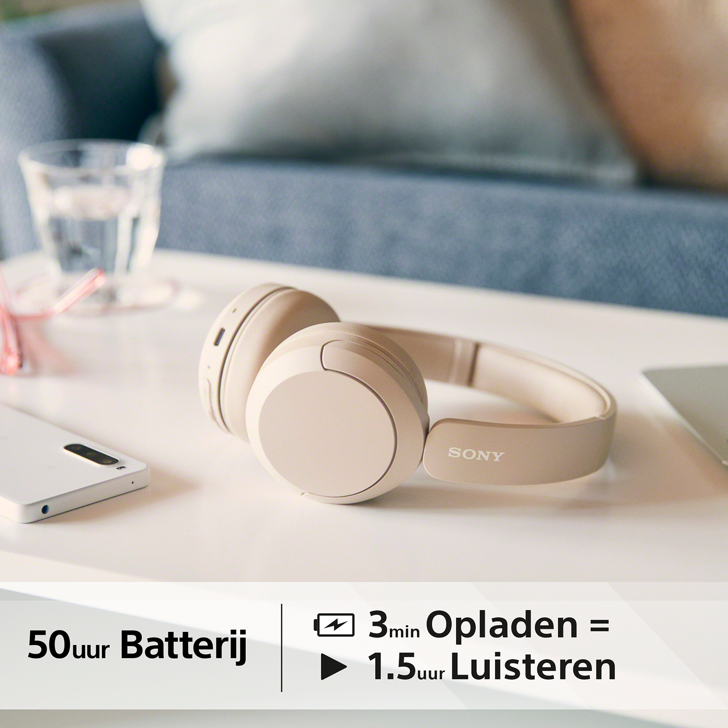 Beige Sony-koptelefoon op een tafel met een telefoon, glas en tekst.