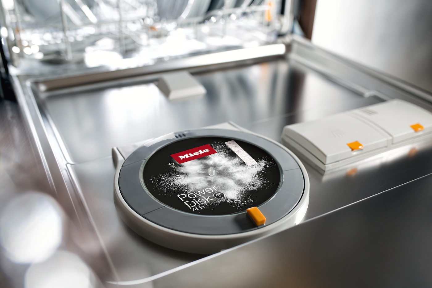 Eine Miele PowerDisk in einem Geschirrspüler. Die Scheibe ist schwarz mit weißem Pulver. Ein rotes Miele-Logo ist vorhanden. Der Geschirrspüler ist silber.