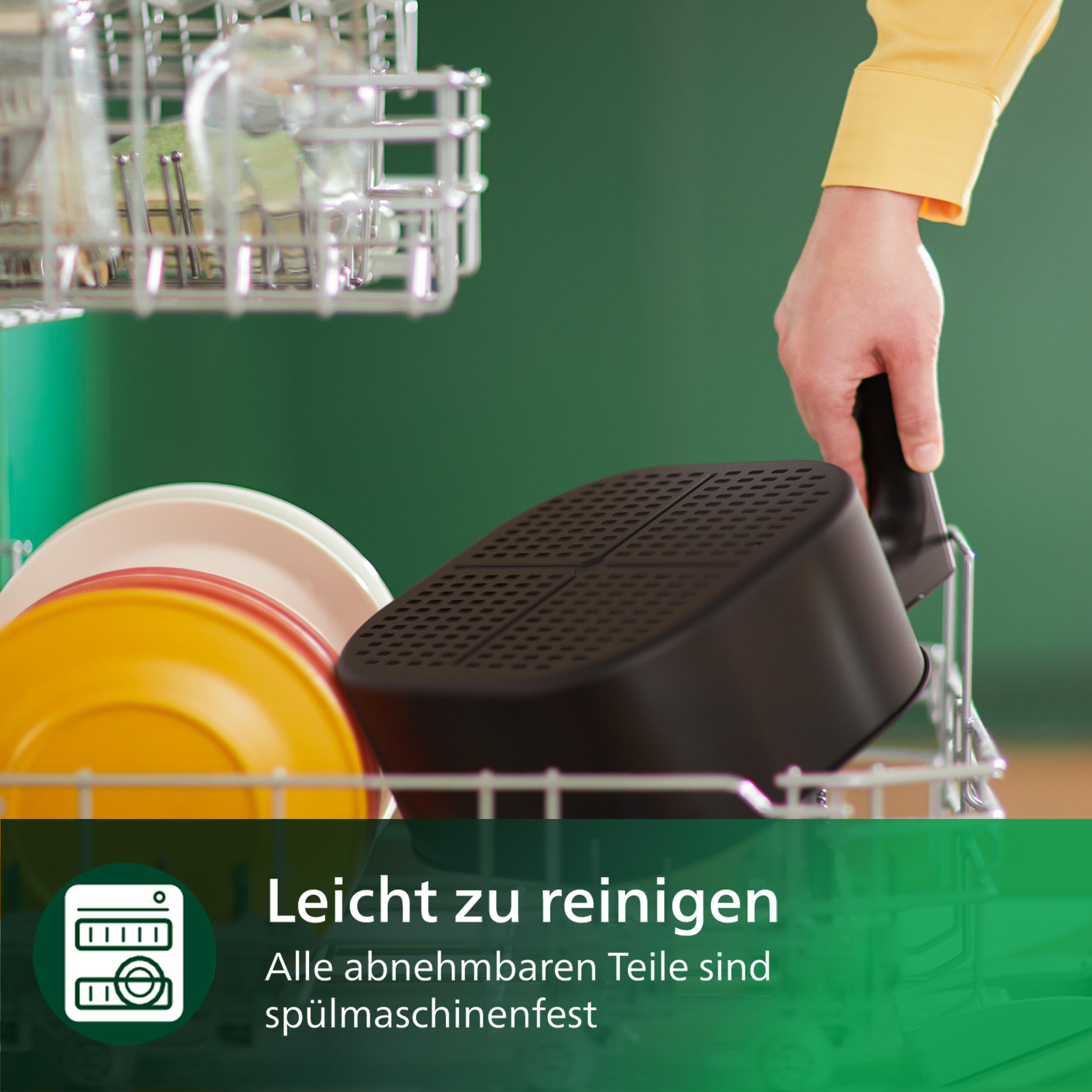 Eine Person nimmt einen schwarzen Airfryer-Korb aus einem Geschirrkorb. Gelbe und weiße Teller sichtbar.