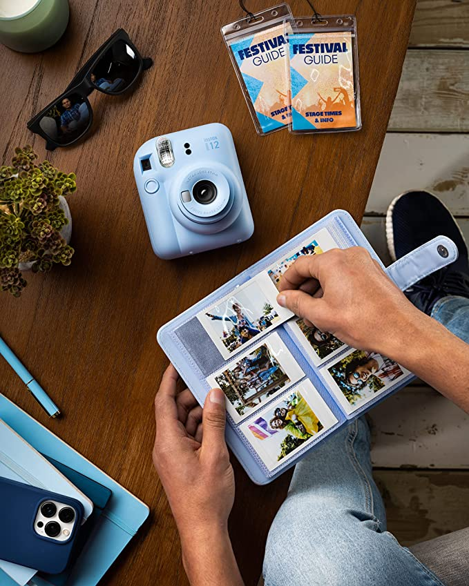 Una persona mira fotos en un álbum de fotos con una cámara Instax azul, gafas de sol y un bolígrafo sobre una mesa.