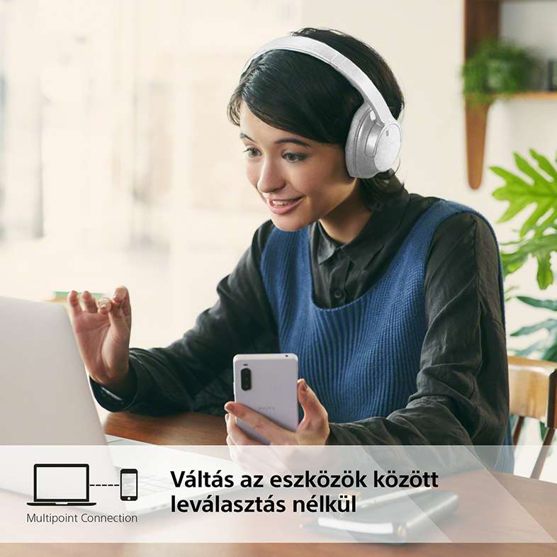 Nő fehér fejhallgatóval, laptopot és telefont használ, mosolyog.