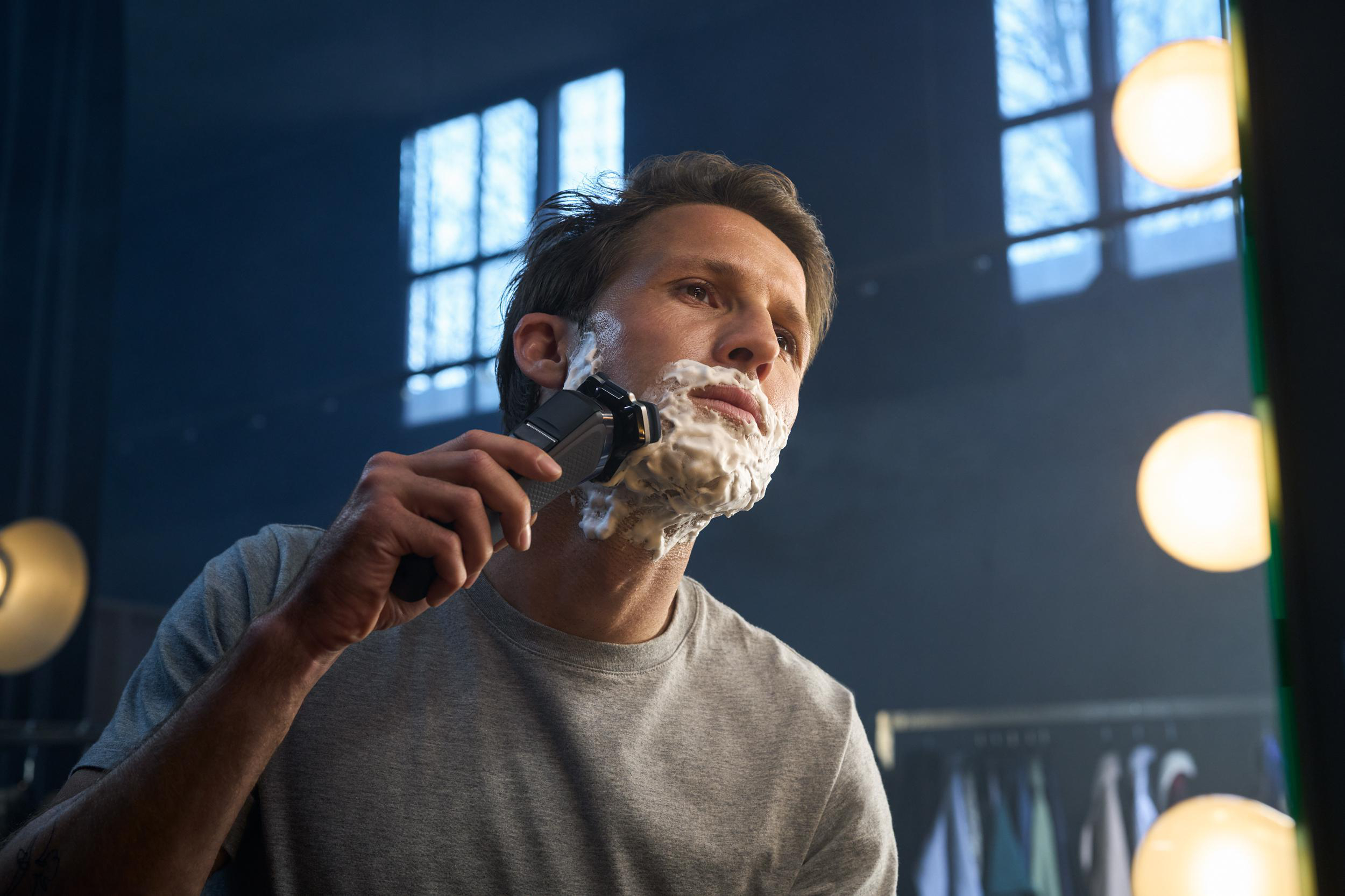 Mann rasiert sich mit Elektrorasierer. Spiegel reflektiert Hintergrund. Er hat Rasierschaum im Gesicht.