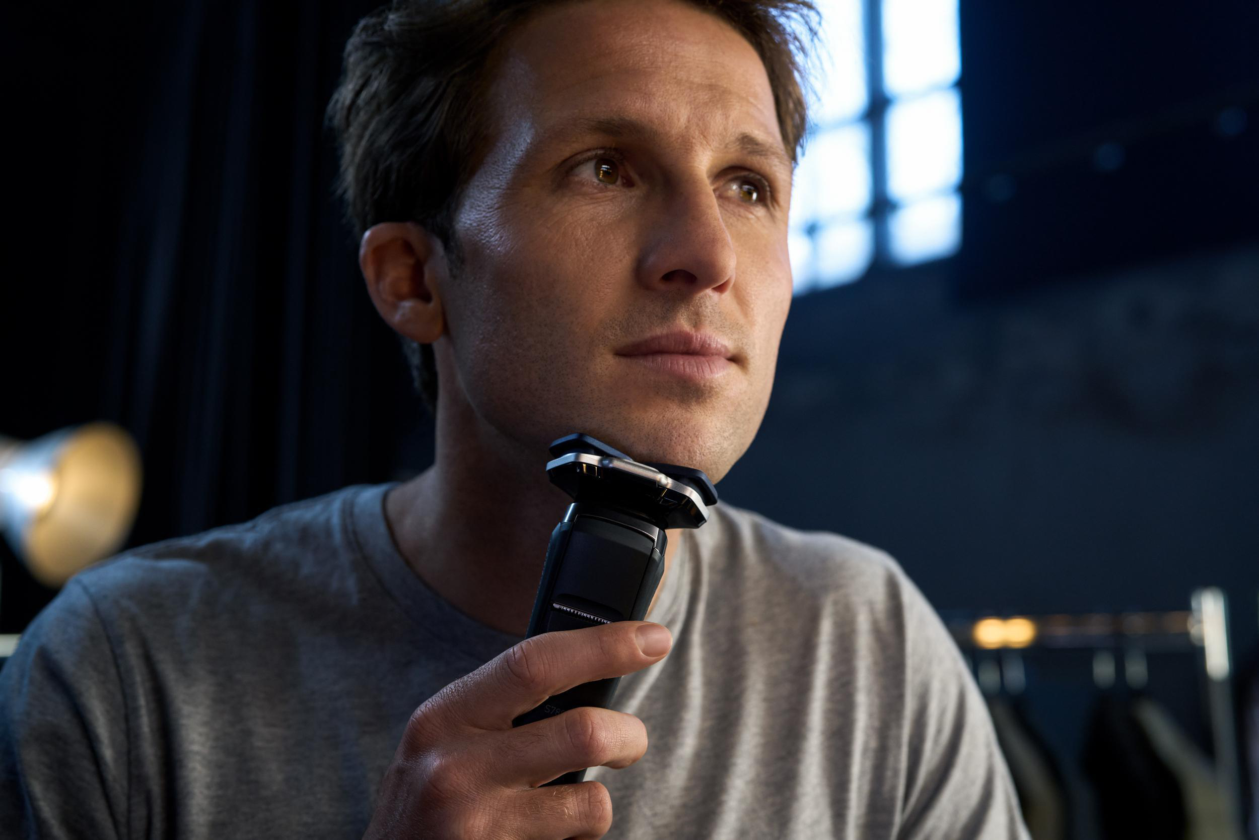 Mann rasiert sich mit einem schwarz-silbernen Rasierer. Er trägt ein graues Hemd. Der Hintergrund ist dunkel.