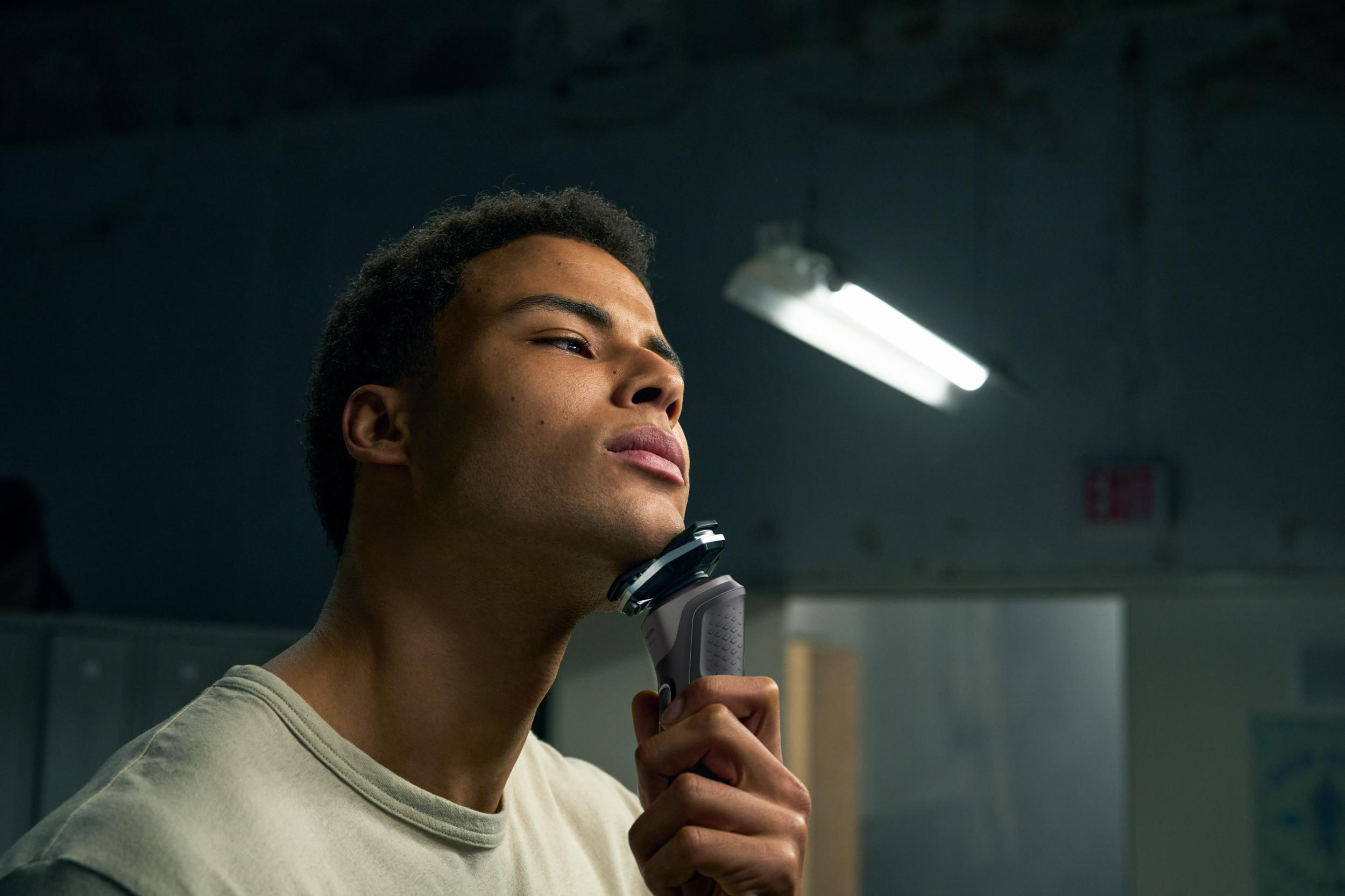 Mann rasiert sich mit einem Elektrorasierer. Er hat kurze schwarze Haare und ist in einem schwach beleuchteten Raum.