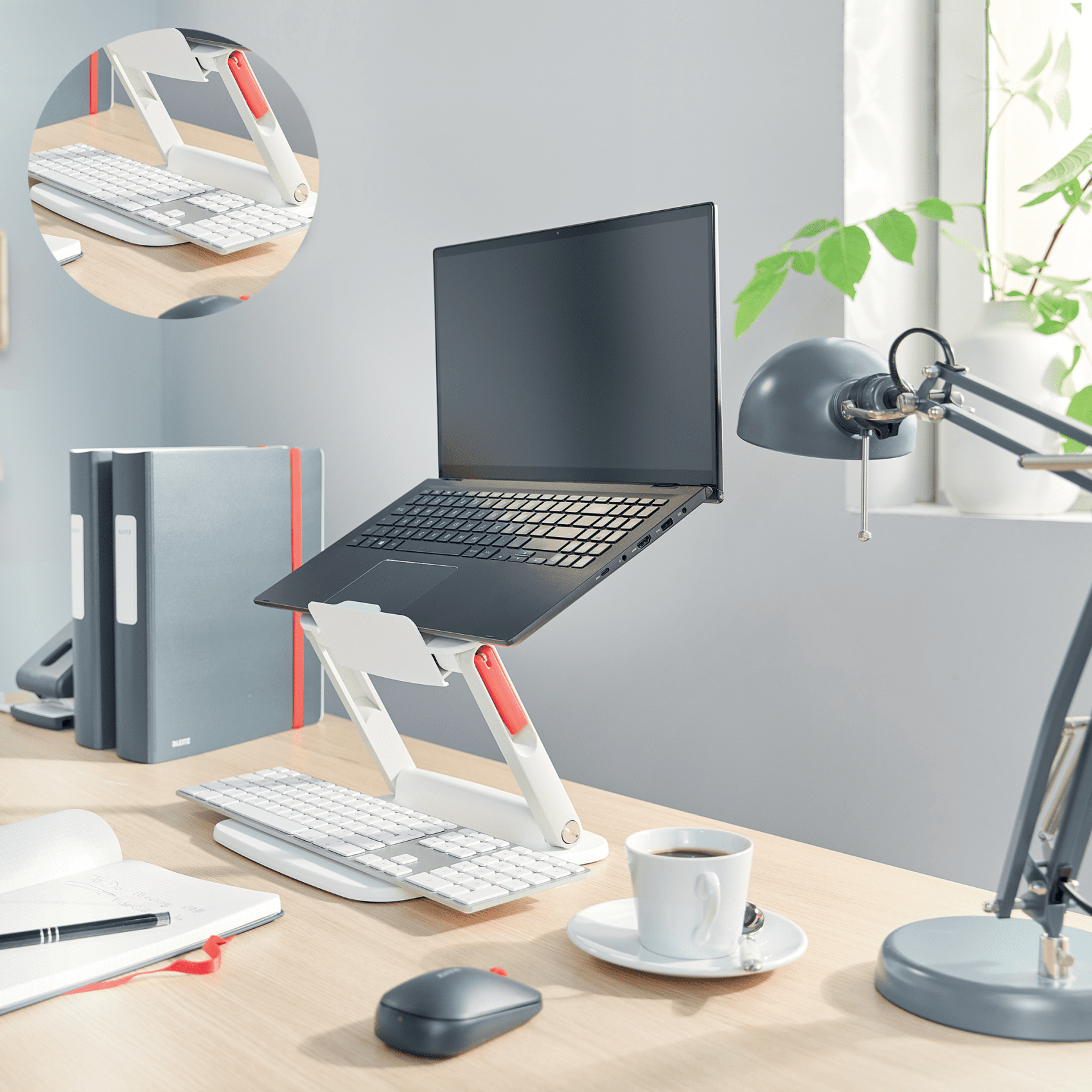 Laptop állványon, billentyűzet, egér, kávéscsésze és lámpa faasztalon.