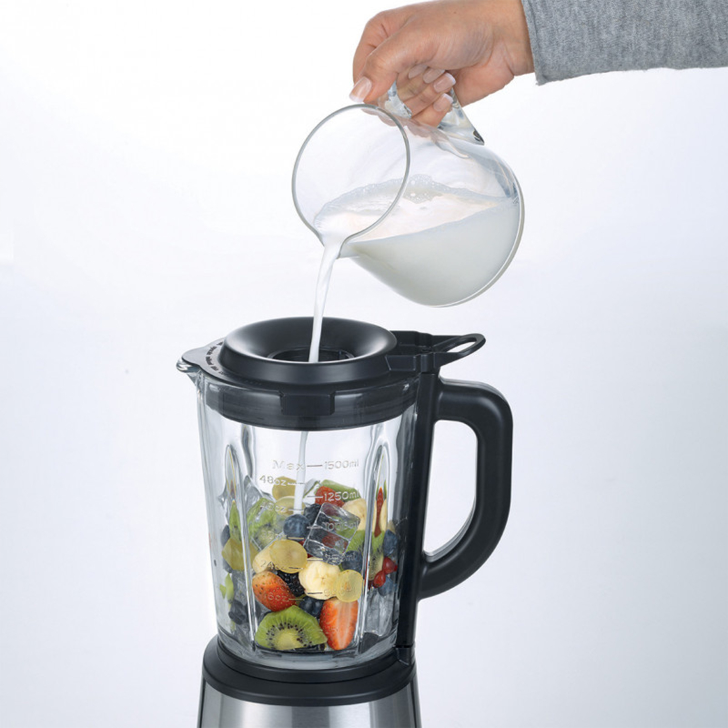 Mano che versa latte da una caraffa di vetro in un frullatore con frutta. Sfondo bianco.