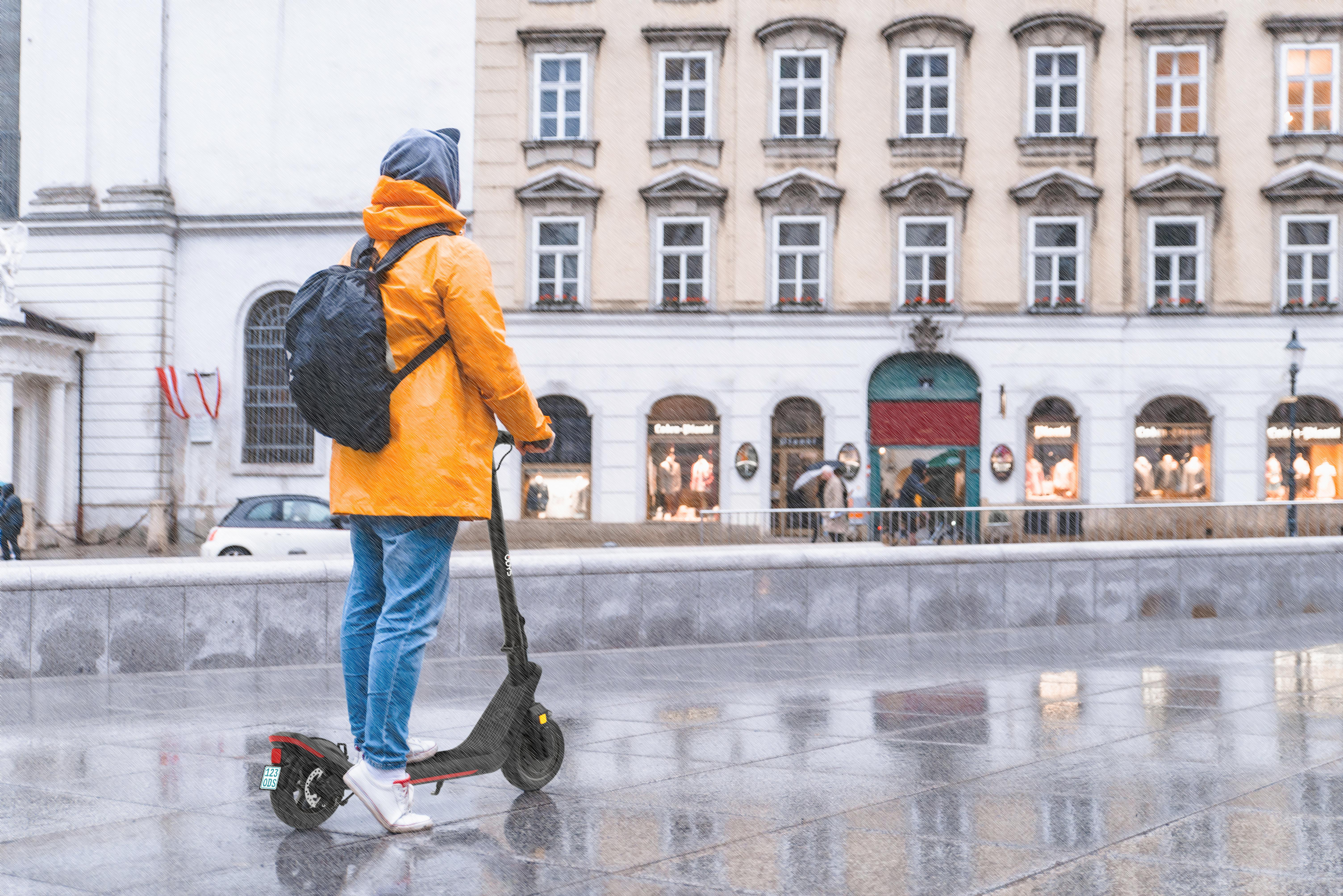 Eine Person in einem orangefarbenen Regenmantel und blauen Jeans, die auf einem Roller auf einer nassen Straße steht.