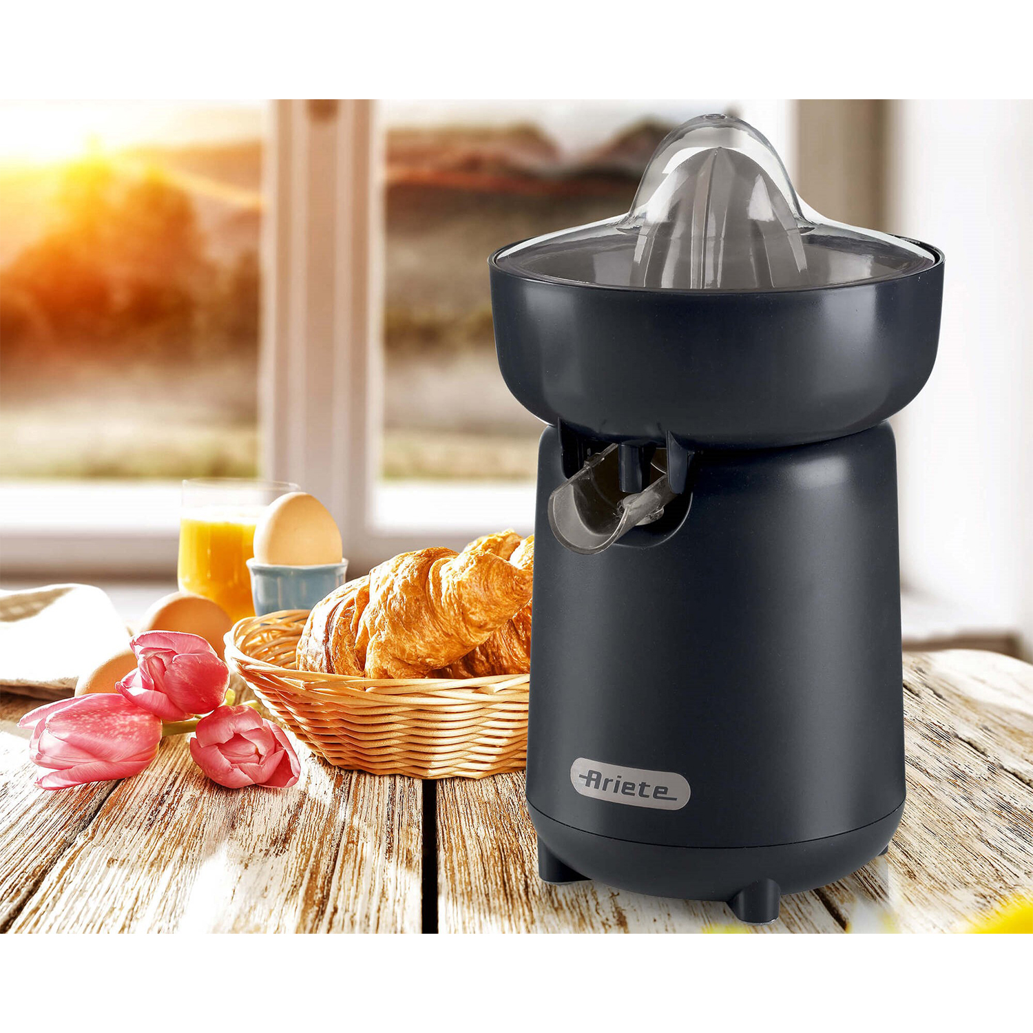 Spremiagrumi elettrico nero con croissant, uova e tulipani su un tavolo di legno vicino a una finestra.