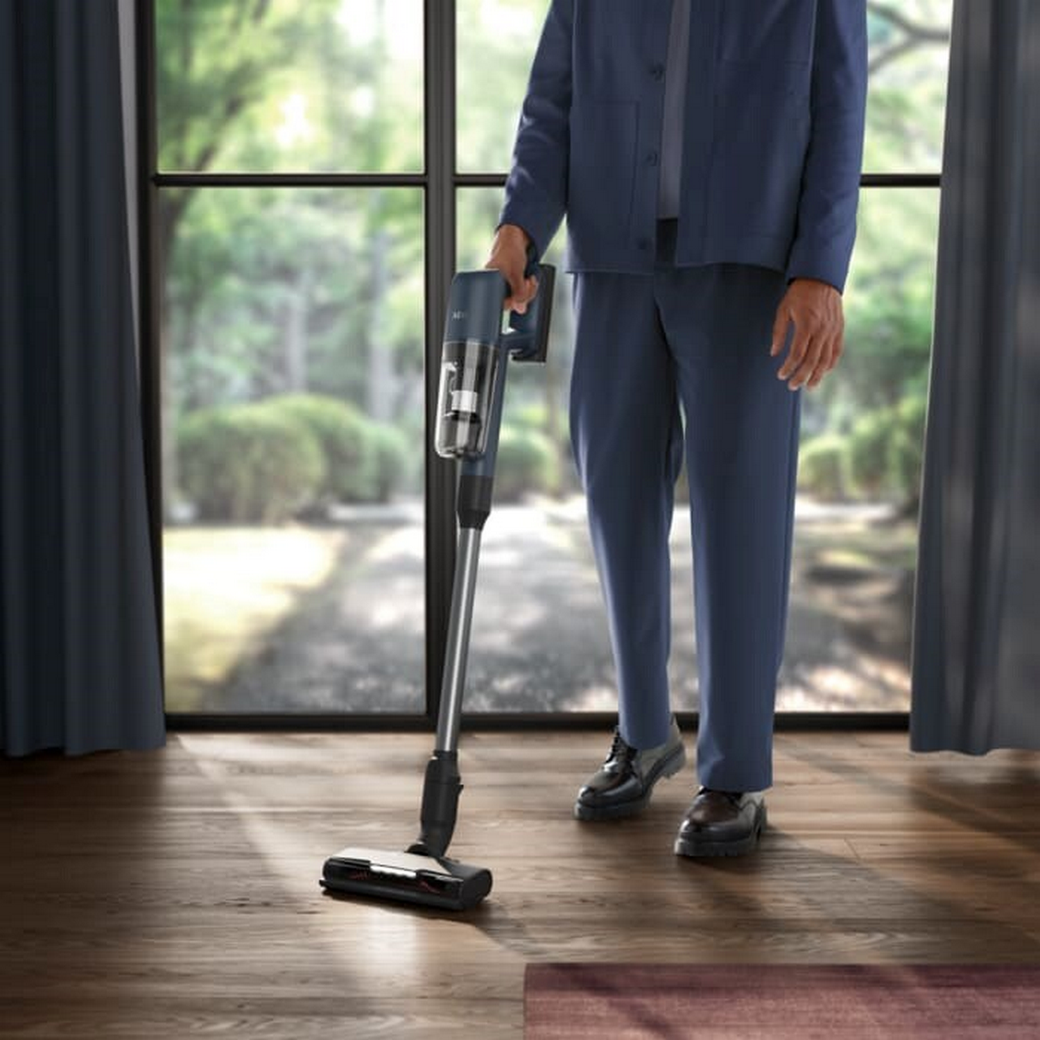 A man in a blue suit vacuums a wooden floor. A window and curtains are in the background.