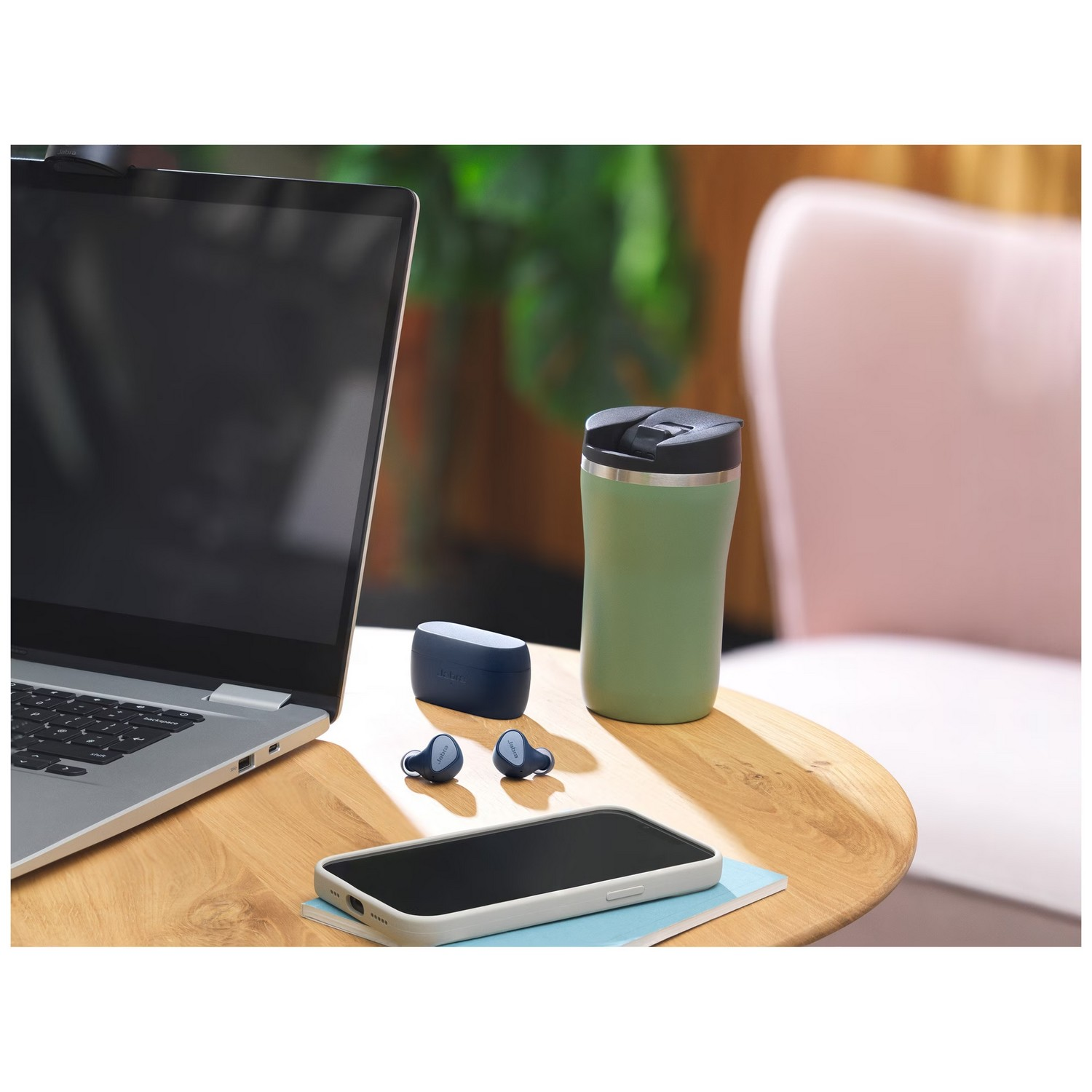 Ordinateur portable, écouteurs, gobelet isotherme et téléphone sur une table en bois. Chaise rose.