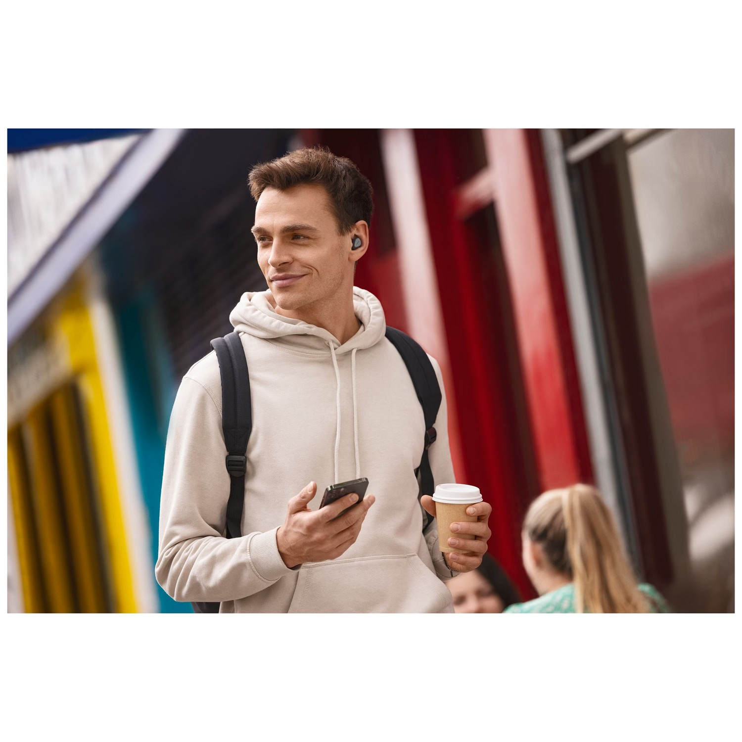 Un homme portant des écouteurs, un sac à dos et tenant un téléphone et une tasse de café.