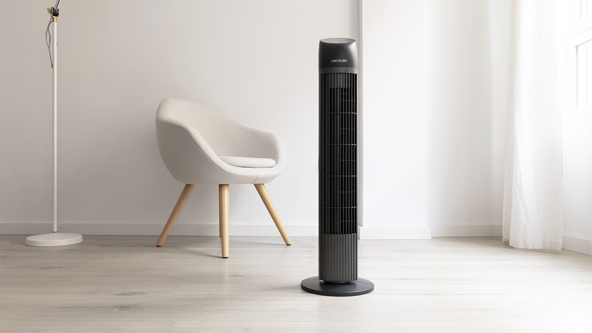 Un ventilador de torre negro en un piso de madera. Paredes blancas, una silla y una lámpara de pie.