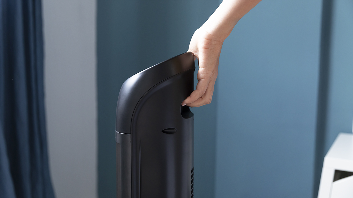 Una mano sosteniendo el asa de un ventilador de torre negro contra una pared azul.