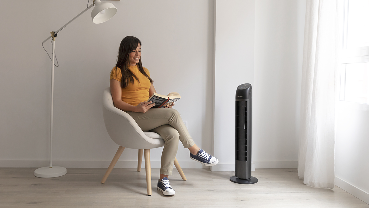 Mujer lee libro, sentada en silla, ventilador al lado. Habitación luminosa, paredes blancas.
