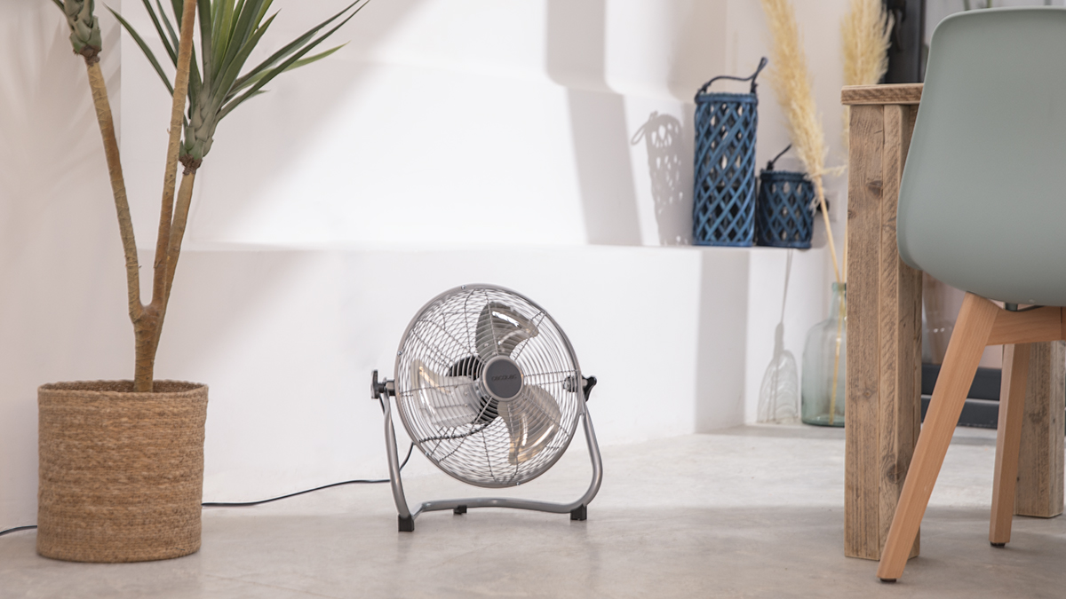 Un ventilador plateado en una habitación con una planta y una mesa, paredes blancas y suelo gris.