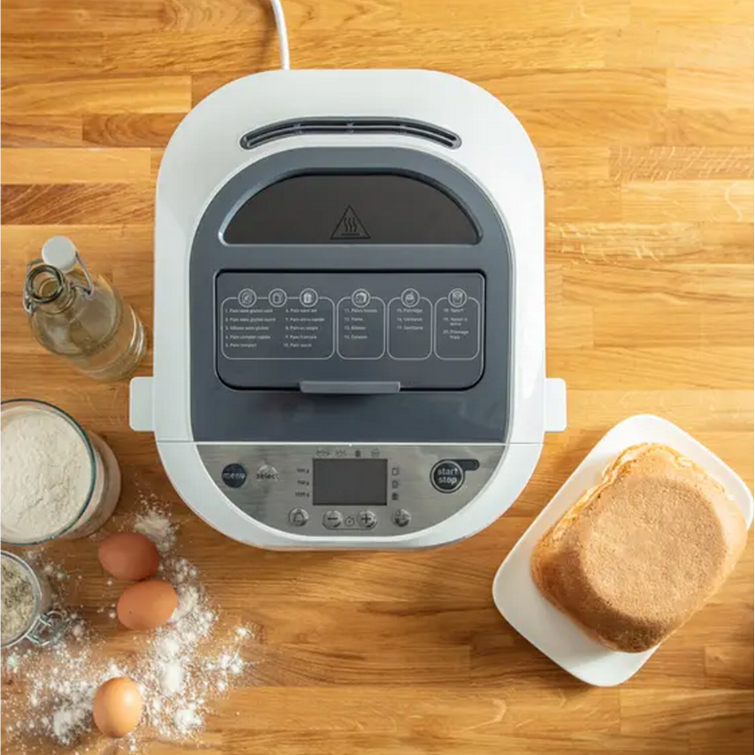 Een broodbakmachine met ingrediënten zoals eieren en bloem eromheen, en een brood aan de zijkant.