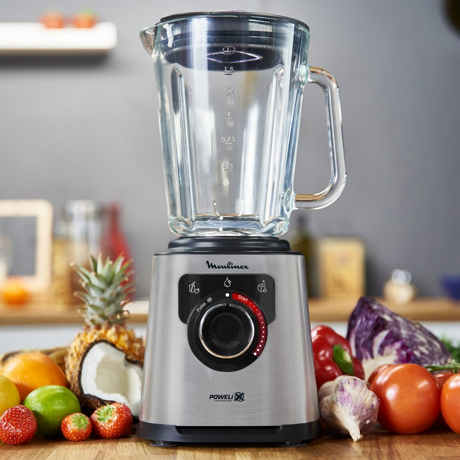 Blender with fruits. A glass blender jar sits on the base. Fruits like pineapple and tomatoes are around.