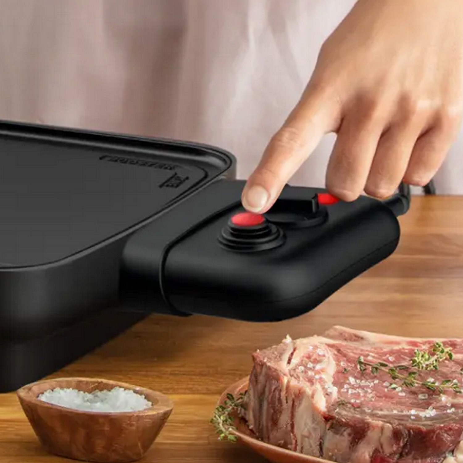 La main d'une personne appuie sur un bouton rouge sur un appareil noir, à côté d'un steak et du sel.