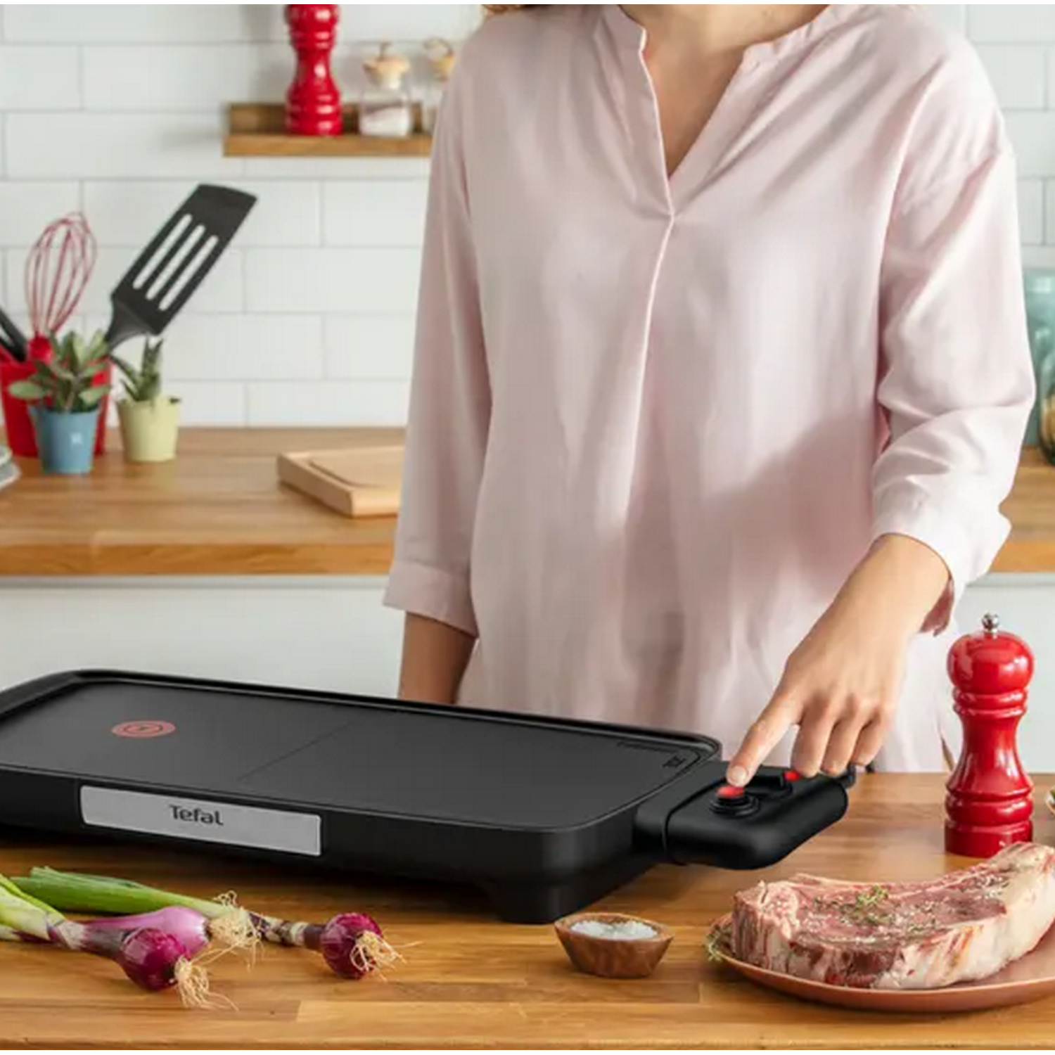 Une femme en chemise rose utilise une plancha noire avec steak et légumes.