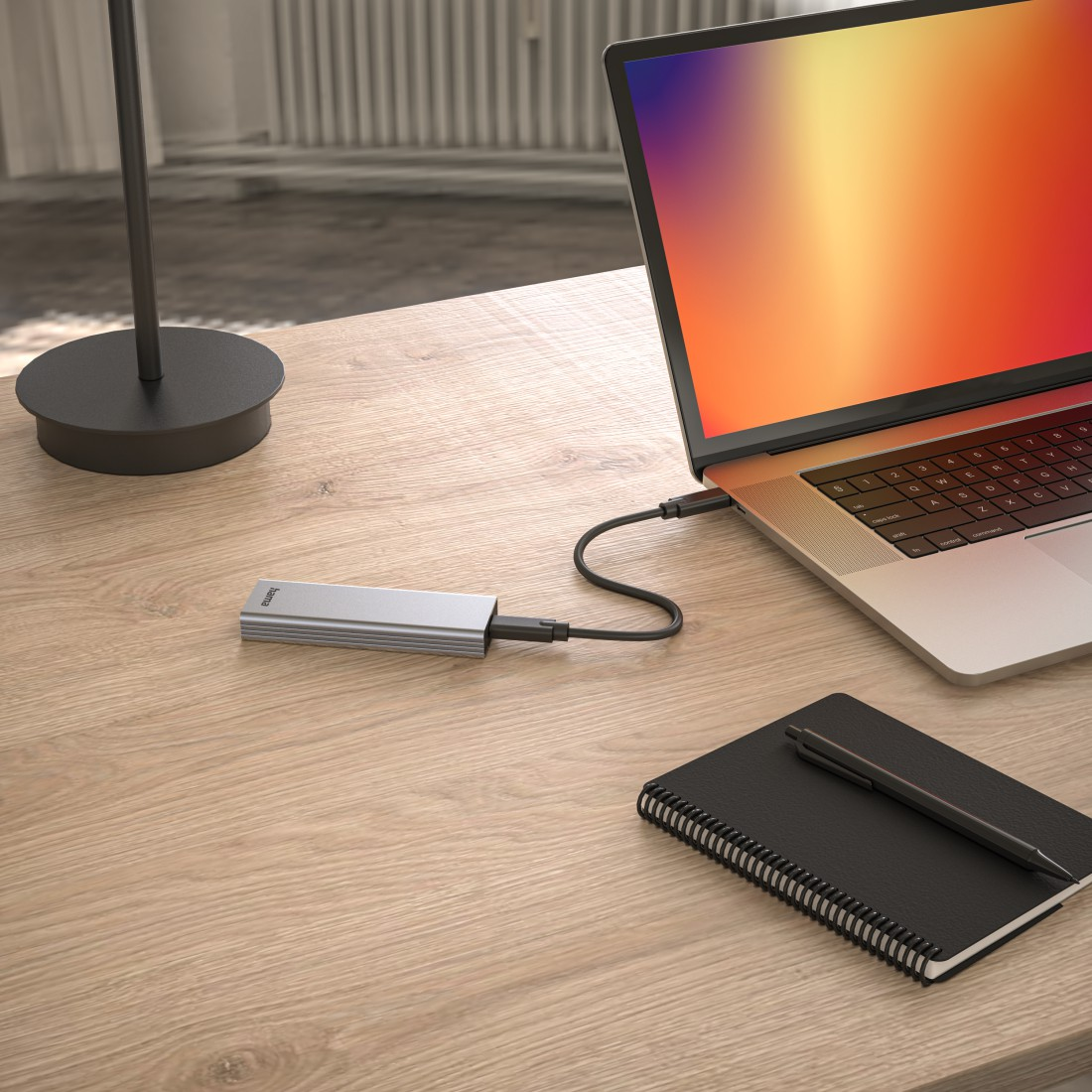 Laptop, der mit einem silbernen Hub verbunden ist. Notizbuch und Stift liegen auf dem Holztisch.