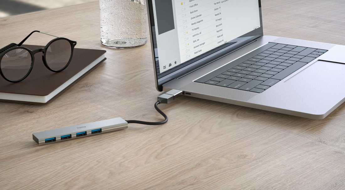 Laptop egy USB-elosztóhoz csatlakoztatva egy fa asztalon, szemüveggel és jegyzetfüzettel.