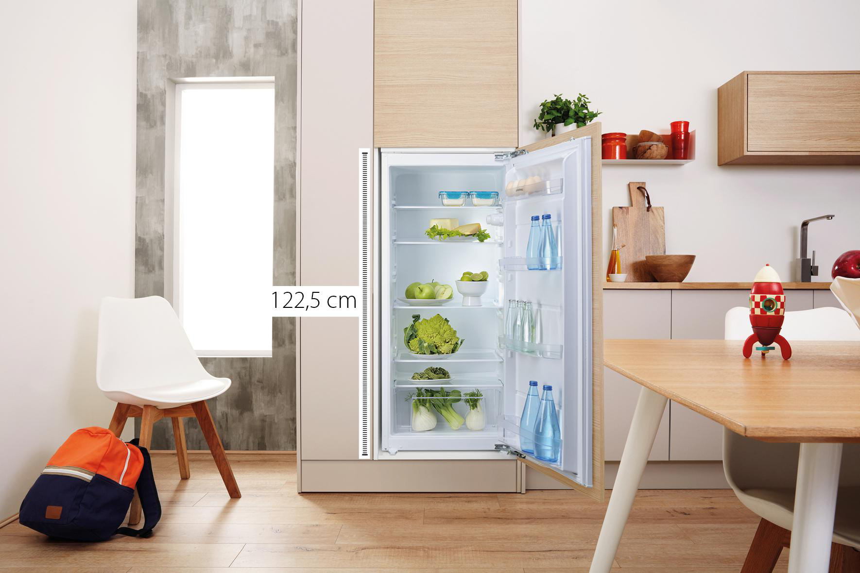 Ein offener Kühlschrank mit Essen darin. Ein weißer Stuhl und ein Rucksack sind im Bild.