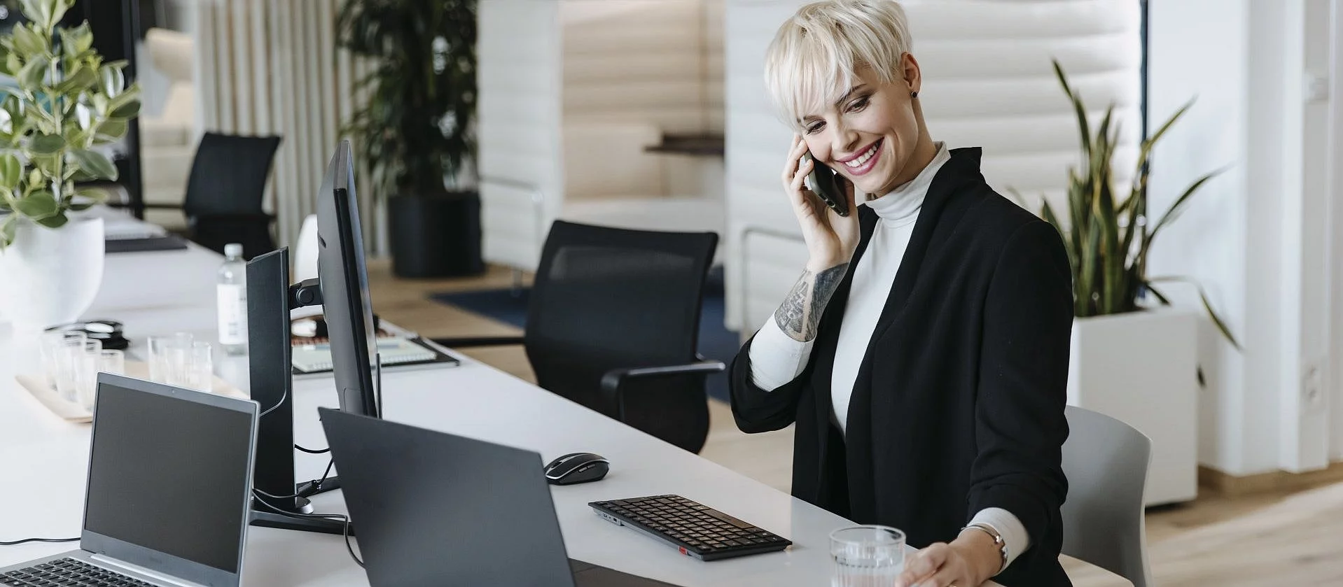 Eine blonde Frau mit kurzer Frisur sitzt in einem modernen Büro und telefoniert glücklich mit ihrem Smartphone.