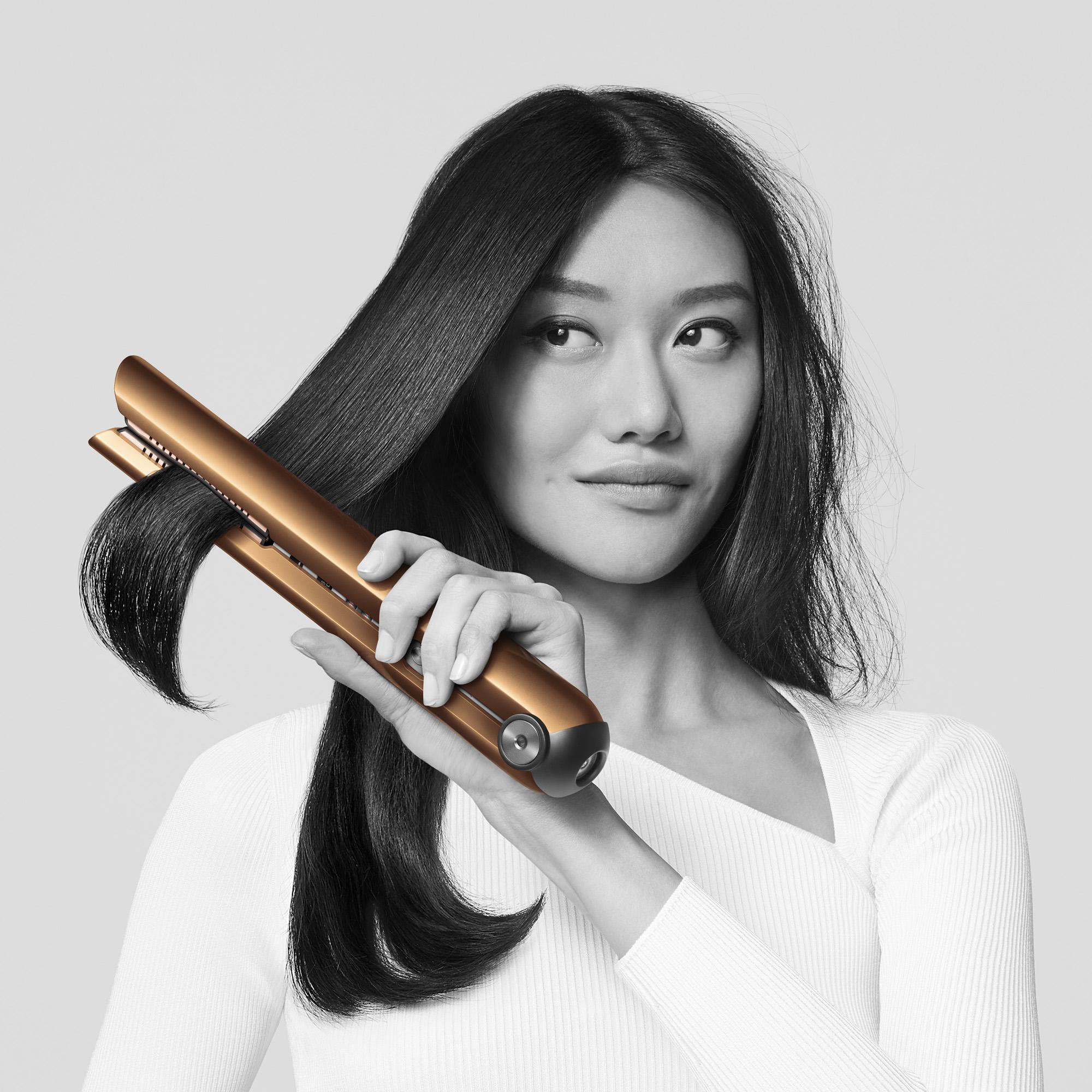 Frau mit glattem Haar benutzt ein goldenes Glätteisen, vor grauem Hintergrund.