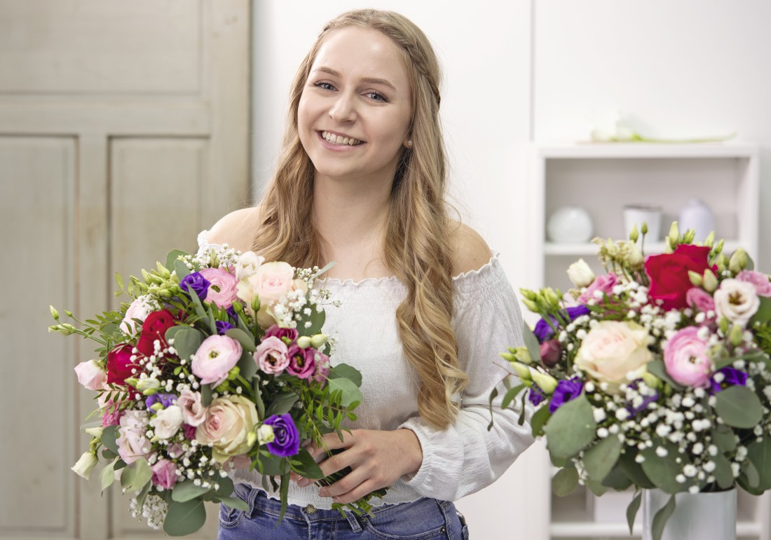 Donna bionda con due bouquet colorati, sorridente. Lo sfondo è una stanza bianca.