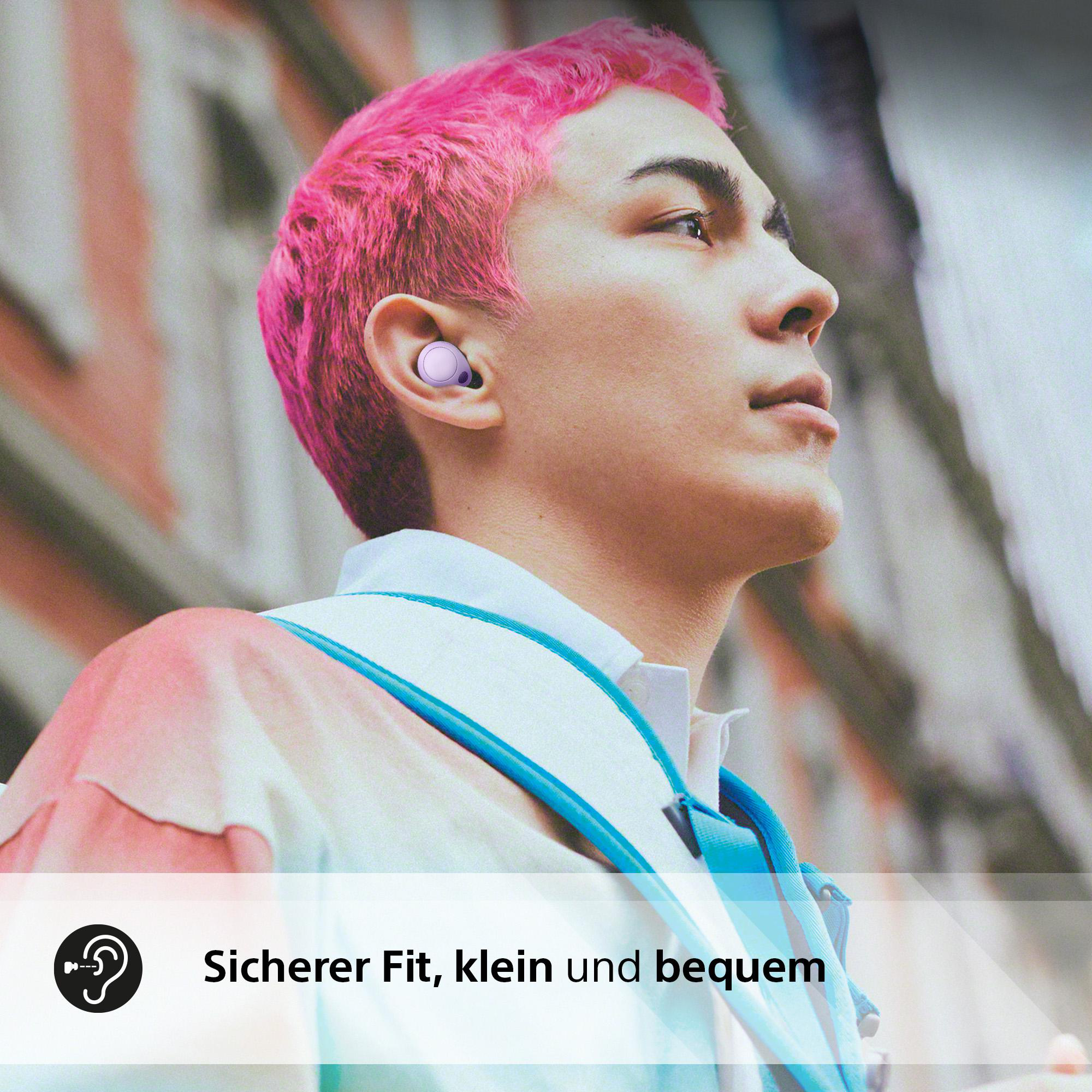 Eine junge Person mit rosa Haaren trägt Ohrhörer. Sie stehen vor einem verschwommenen Hintergrund.