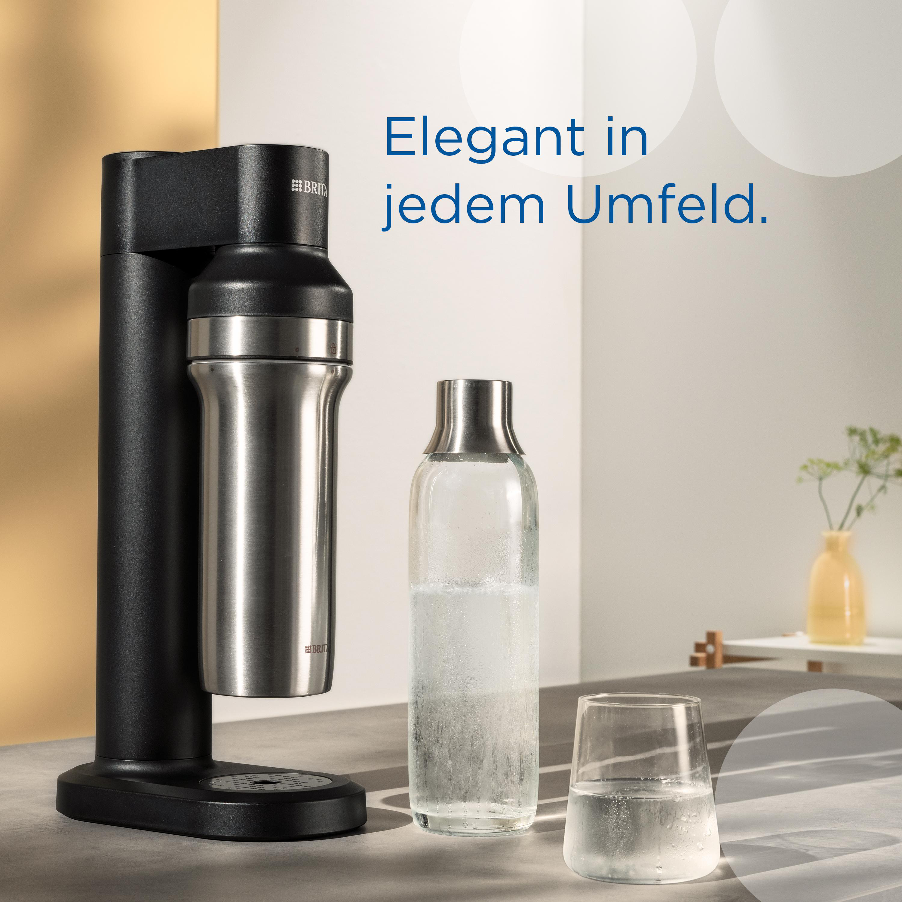 Ein schwarzer Sodabereiter mit einer Edelstahlflasche und einem Glas Wasser stehen auf einem Tisch. Daneben steht auch eine Glasflasche.