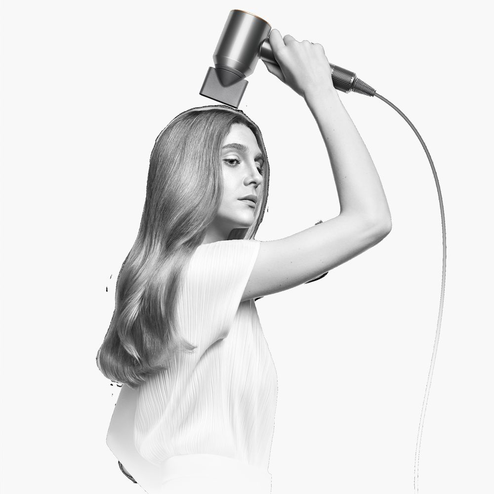 Mujer secándose el pelo con un secador de pelo plateado, inclinado hacia su cabeza. El fondo es blanco.