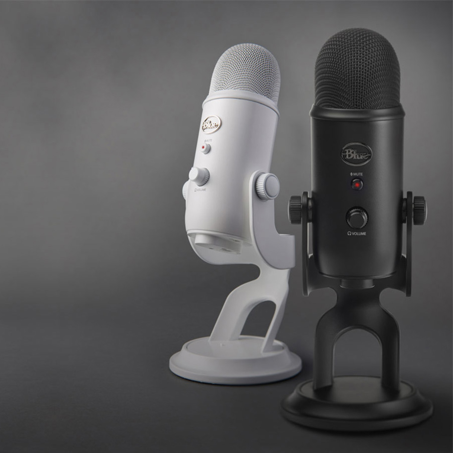 Zwei Blue Yeti Mikrofone, eines weiß, eines schwarz, auf grauem Hintergrund. Das weiße ist links, das schwarze rechts.