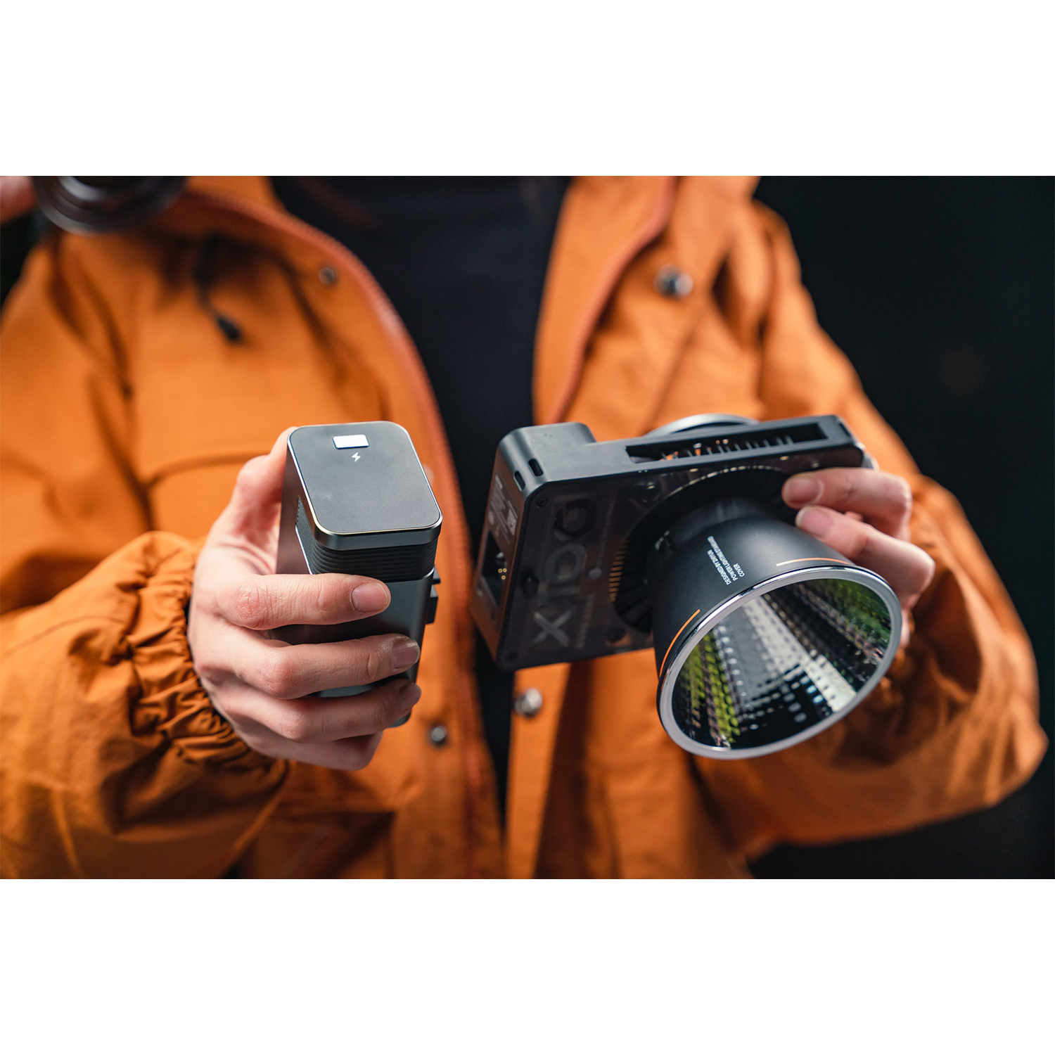Una persona con una giacca arancione tiene in mano una fotocamera con un accessorio luminoso.