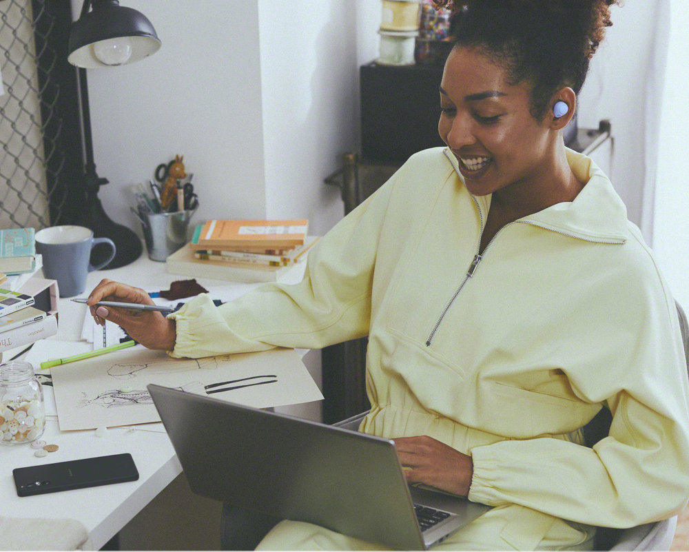 Femme en veste jaune et écouteurs bleus. Elle dessine sur un bureau avec un ordinateur.