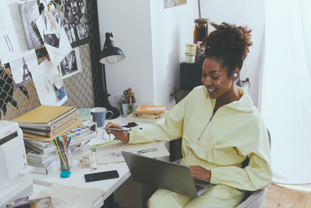 Femme assise à un bureau avec un ordinateur portable et dessinant. Des écouteurs sont dans son oreille.