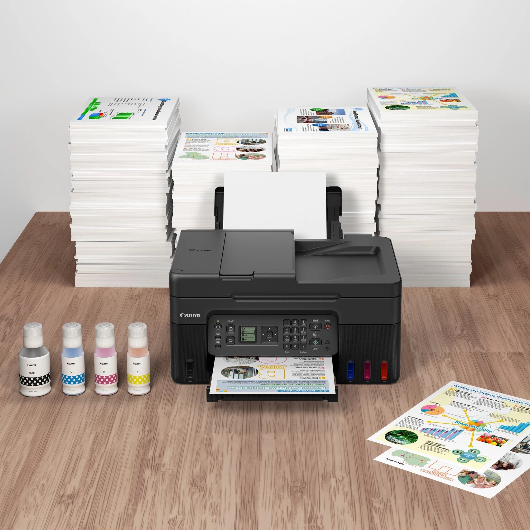 Schwarzer Canon Drucker auf Holztisch mit vier Tintenflaschen und großen Papierstapeln im Hintergrund.