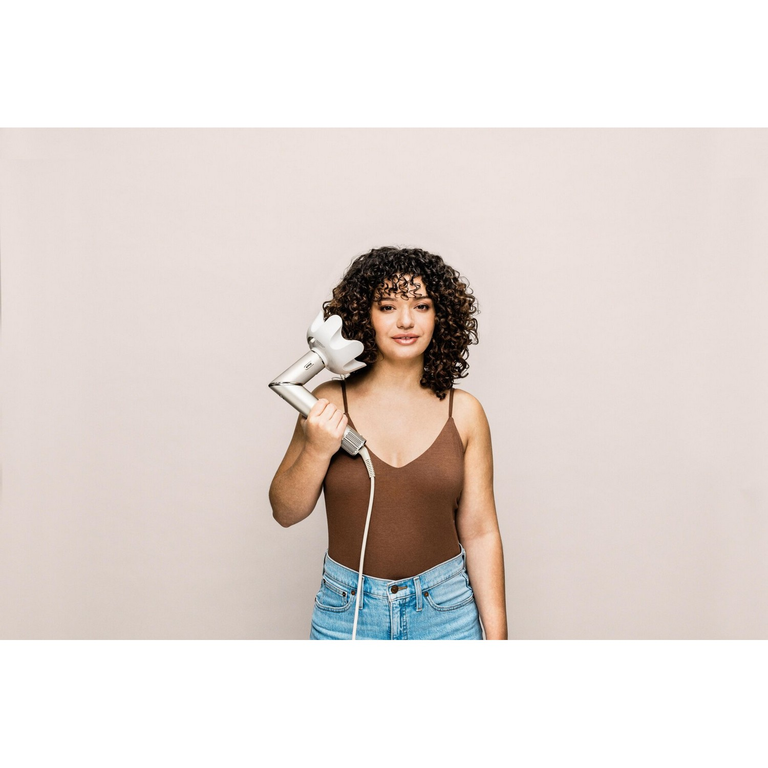 A woman with curly hair holding a white hair dryer, smiling at the camera, against a neutral background.