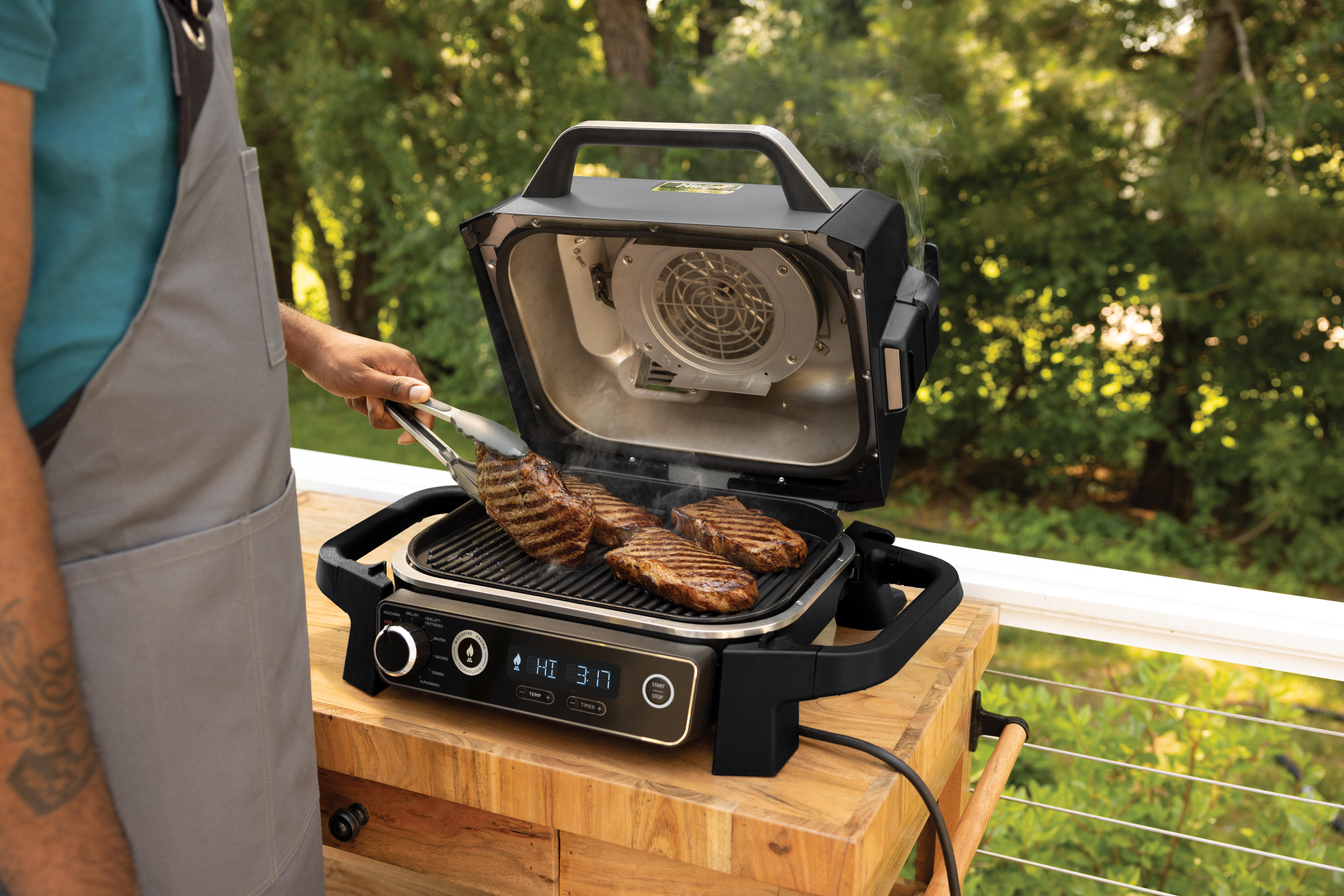 Mann grillt Steaks auf einem Außengrill. Grüne Bäume sind im Hintergrund.